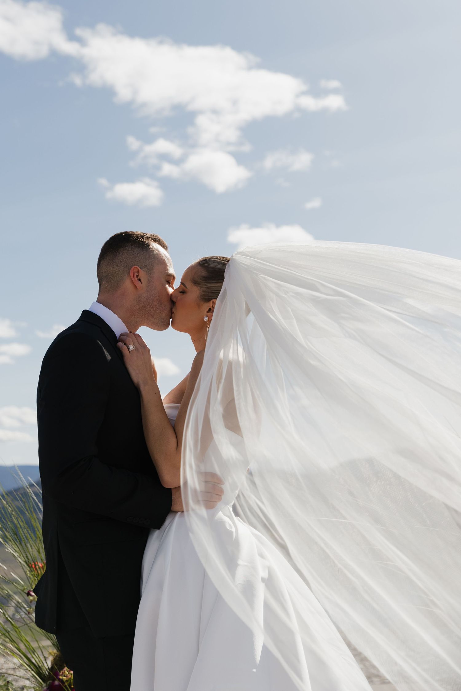 Bride and groom share a romantic kiss outdoors with brides veil billowing in the breeze.