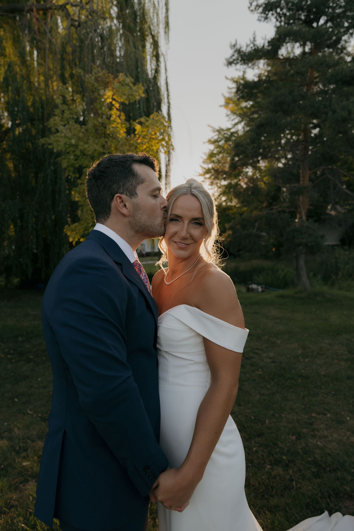 Groom in navy suit kisses bride's cheek at golden hour in a lush green garden setting.