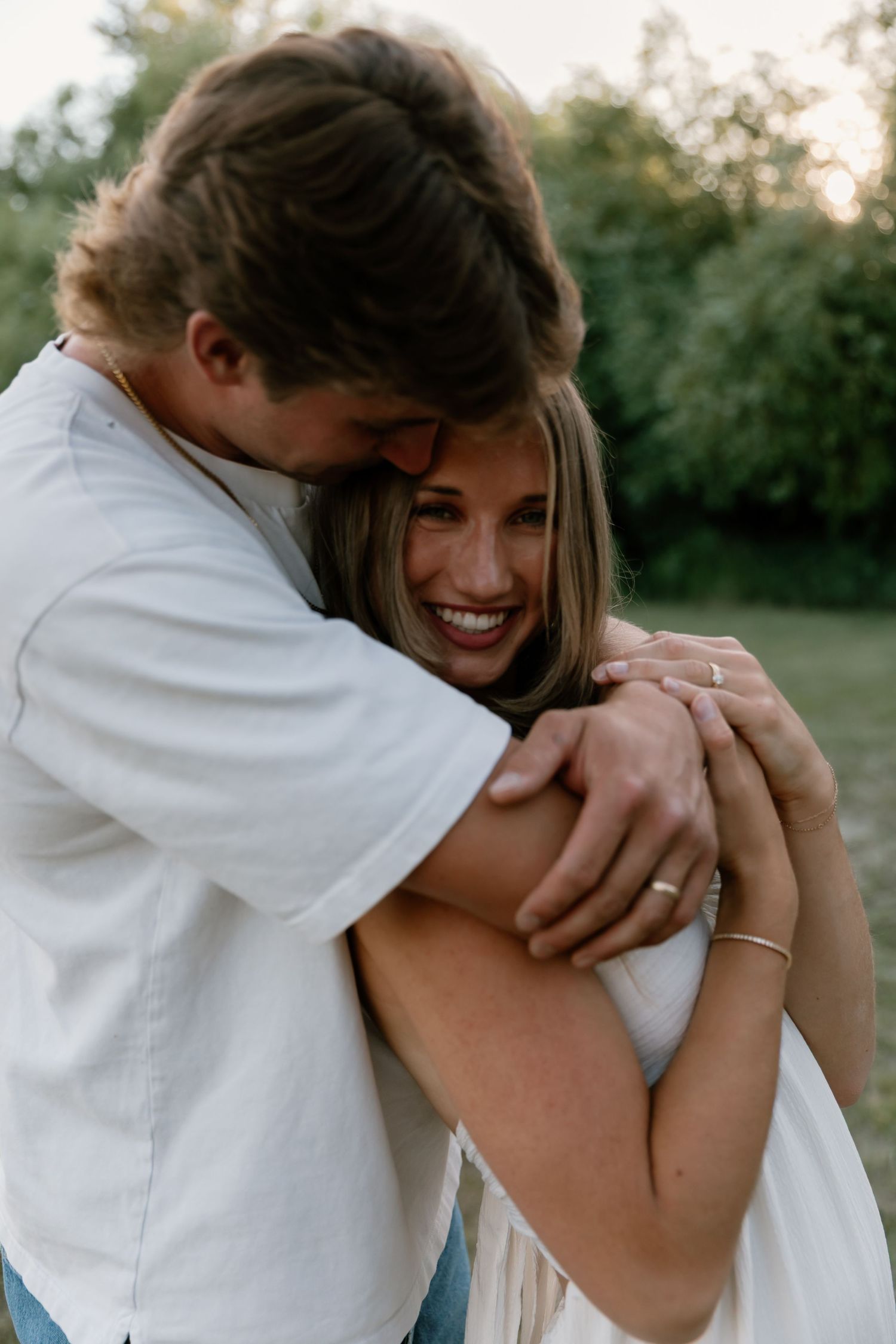 A man hugging a laughing woman from behind in a warm outdoor embrace at golden hour.