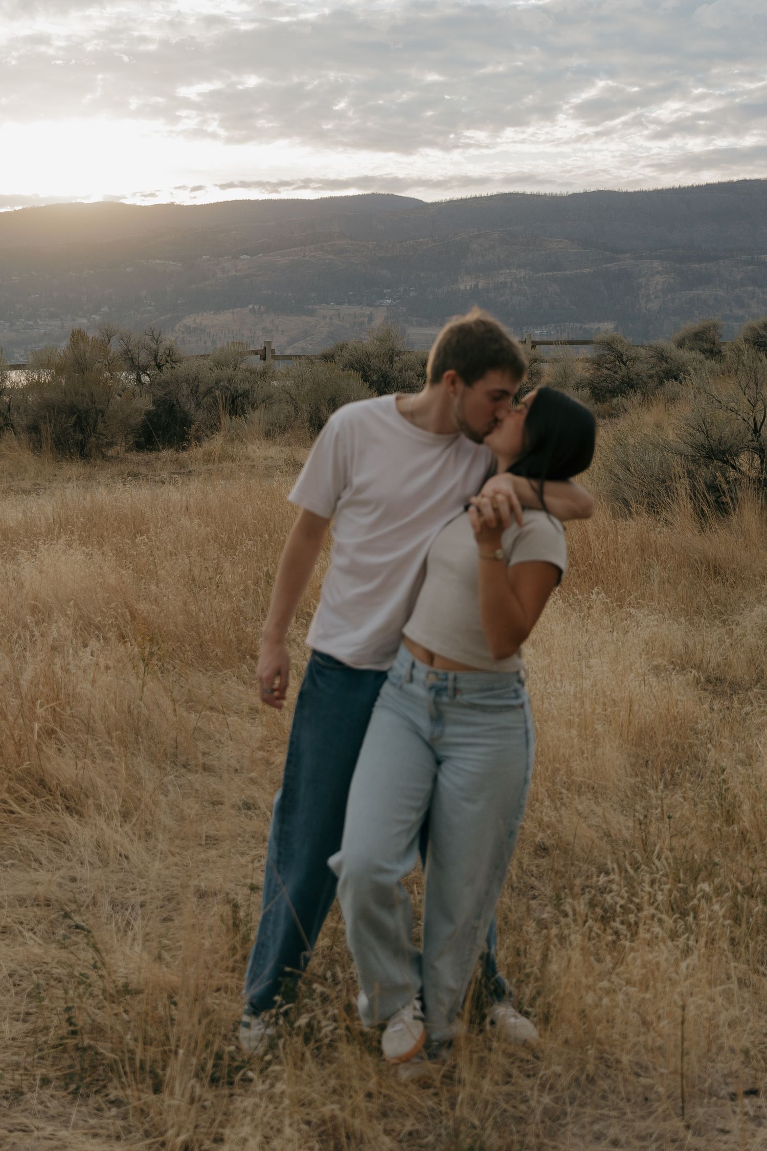 A couple sharing a romantic kiss in a golden field with scenic mountain views at sunset.