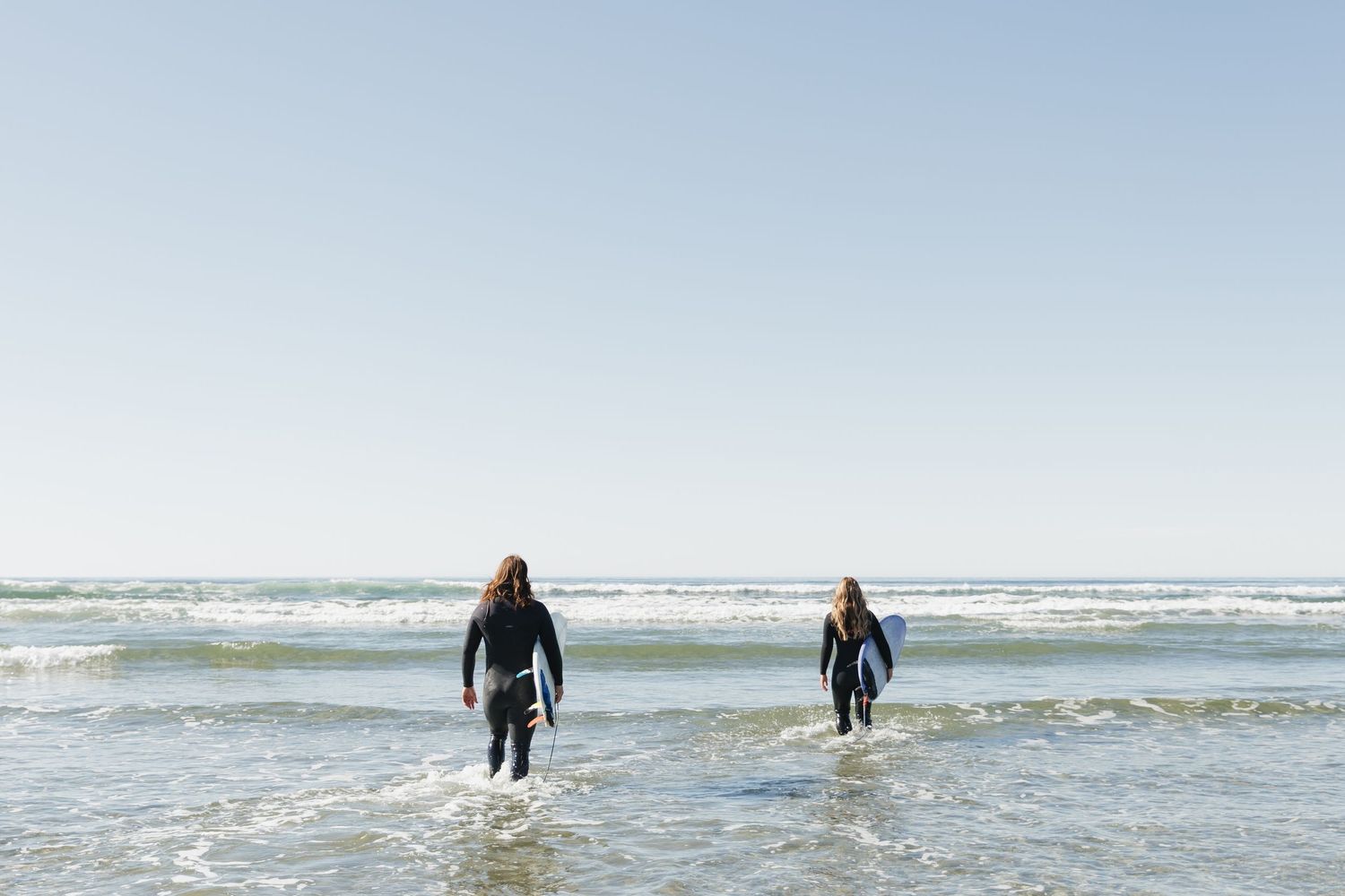 Two surfers in wetsuits walking into ocean waves on a clear sunny beach day.
