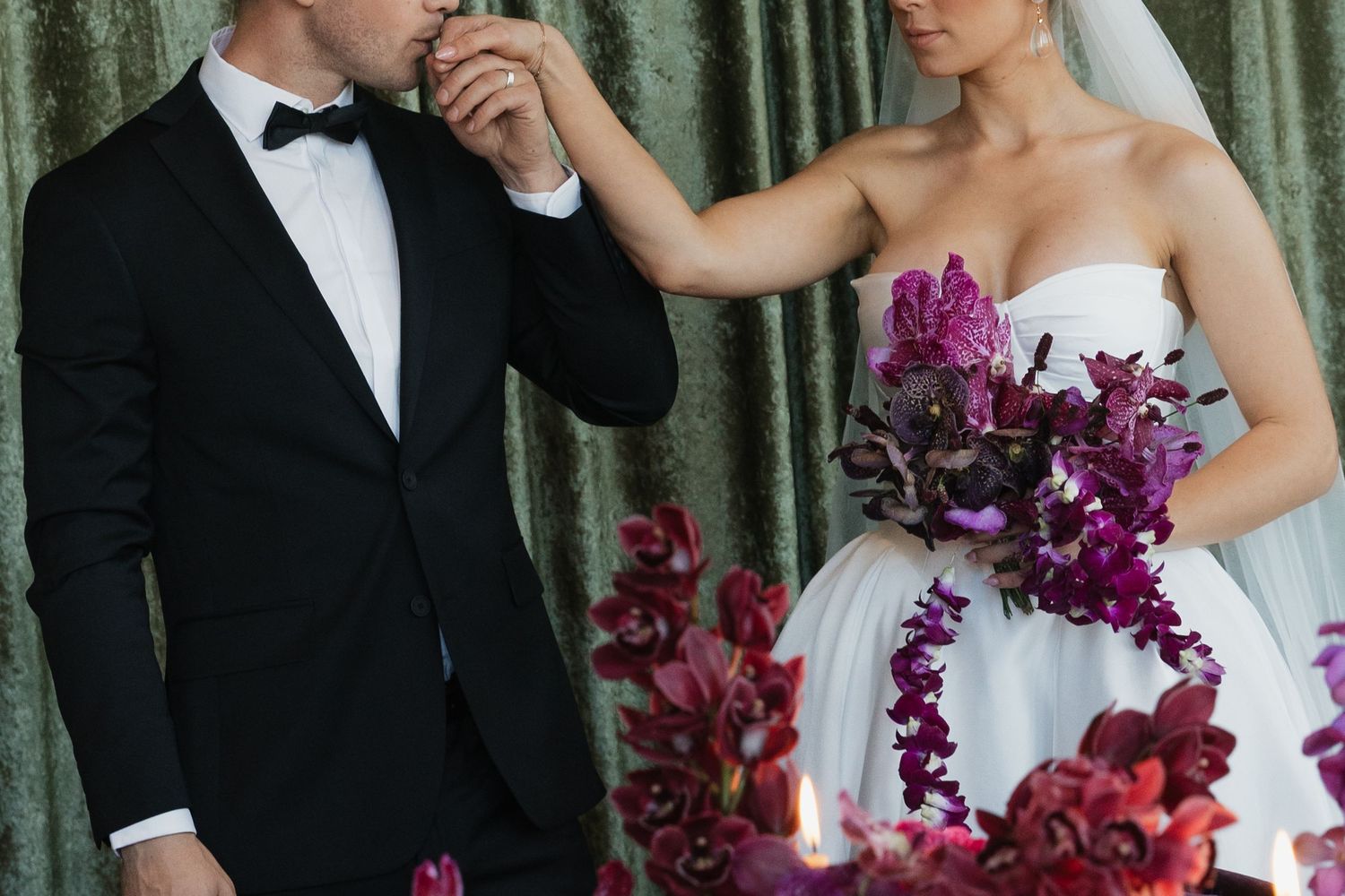 Groom in black tuxedo kisses bride's hand as she holds a lush burgundy orchid bouquet.