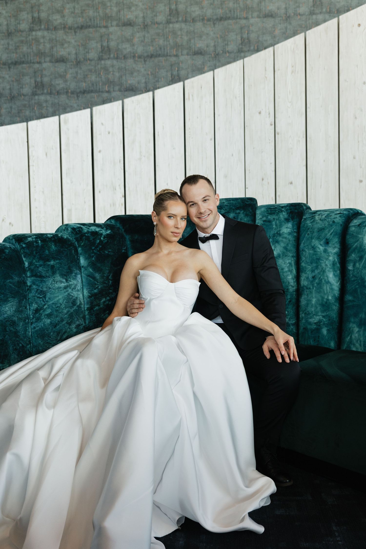 Elegant bride and groom pose on green velvet sofa, bride in strapless ballgown, groom in black tuxedo.
