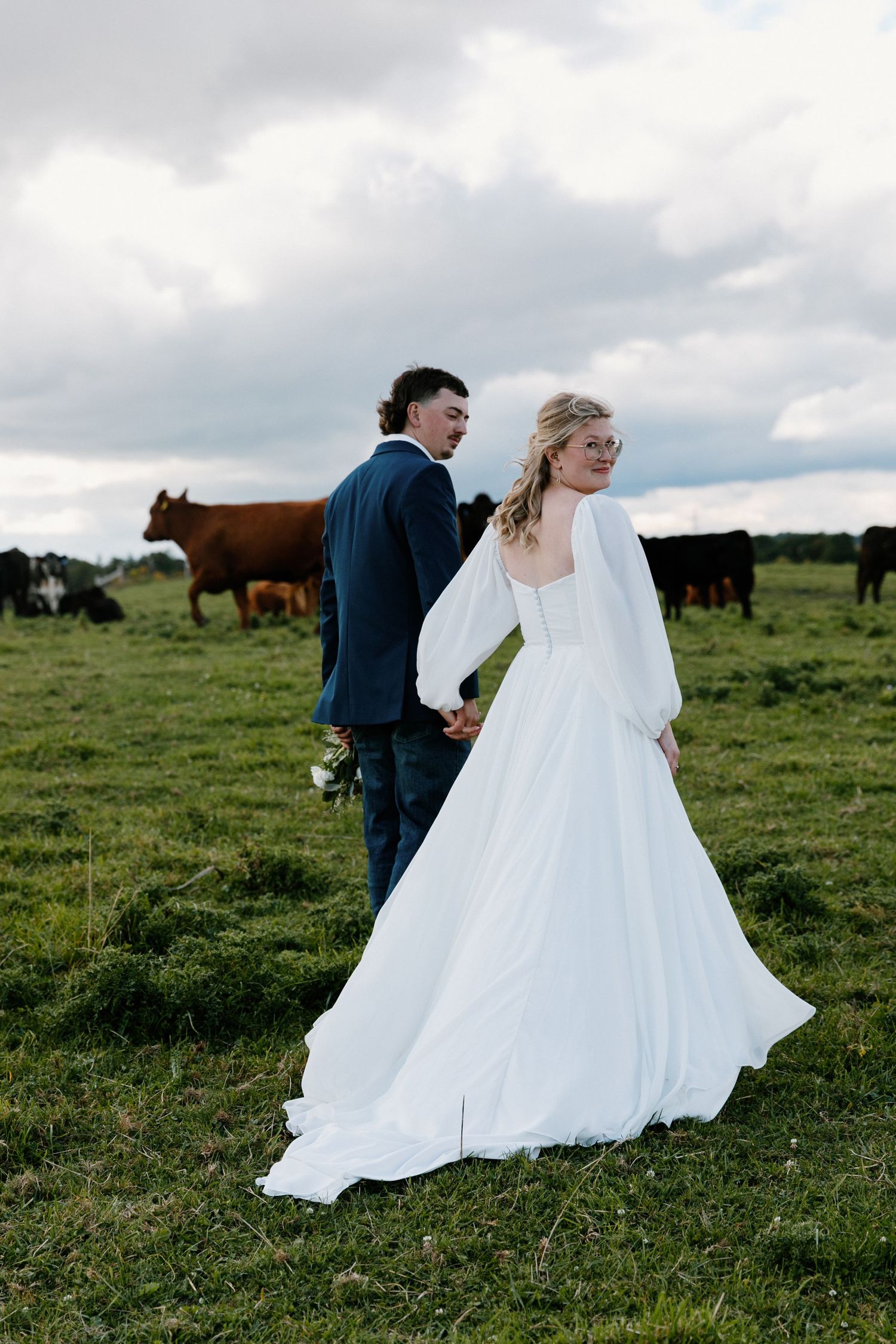 Bride in flowing white gown and groom in navy suit walk hand in hand through a cattle pasture.