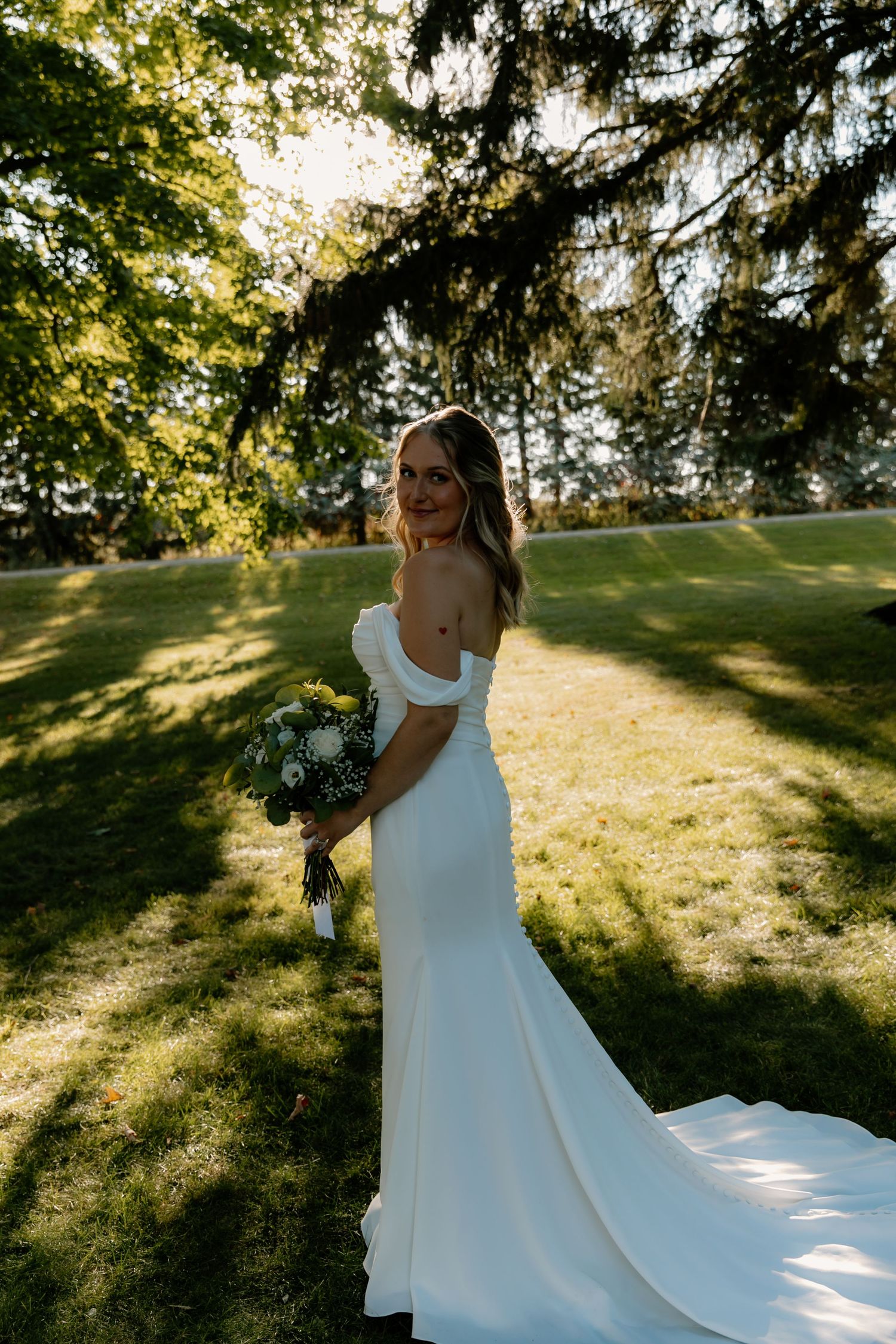 Beautiful bride in fitted off-shoulder wedding gown with train, posing in sunlit park with bouquet.