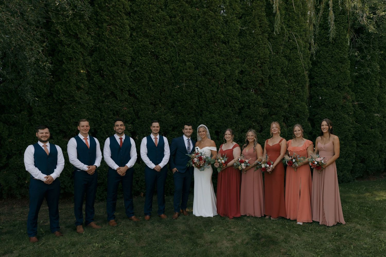 Wedding party poses in front of tall green hedge, bridesmaids in coral and mauve gowns, groomsmen in navy suits.