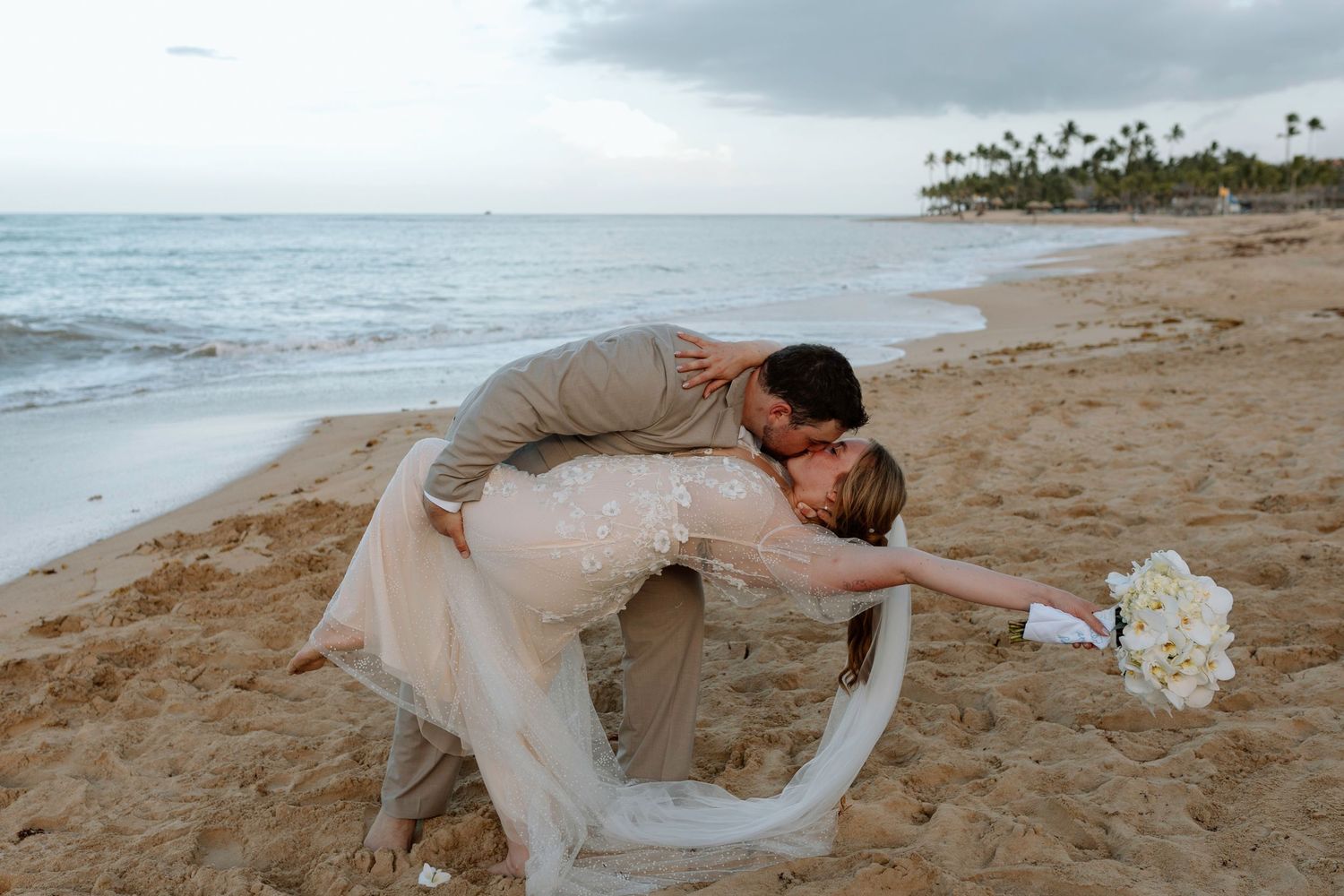 Groom dips bride for romantic kiss on sandy beach holding white flower bouquet.