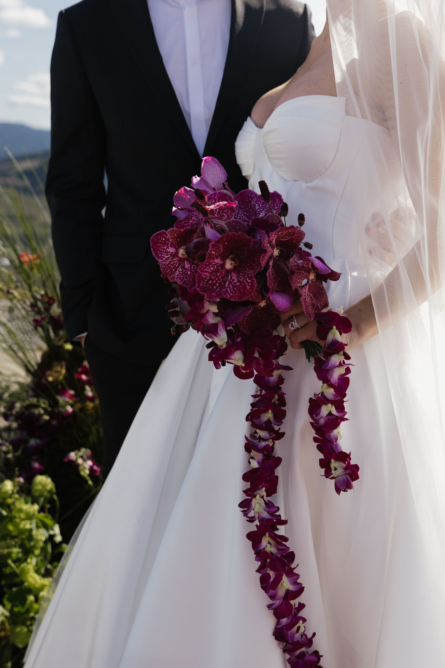 Bride holds a stunning cascading bouquet of deep burgundy orchids against her white wedding gown.