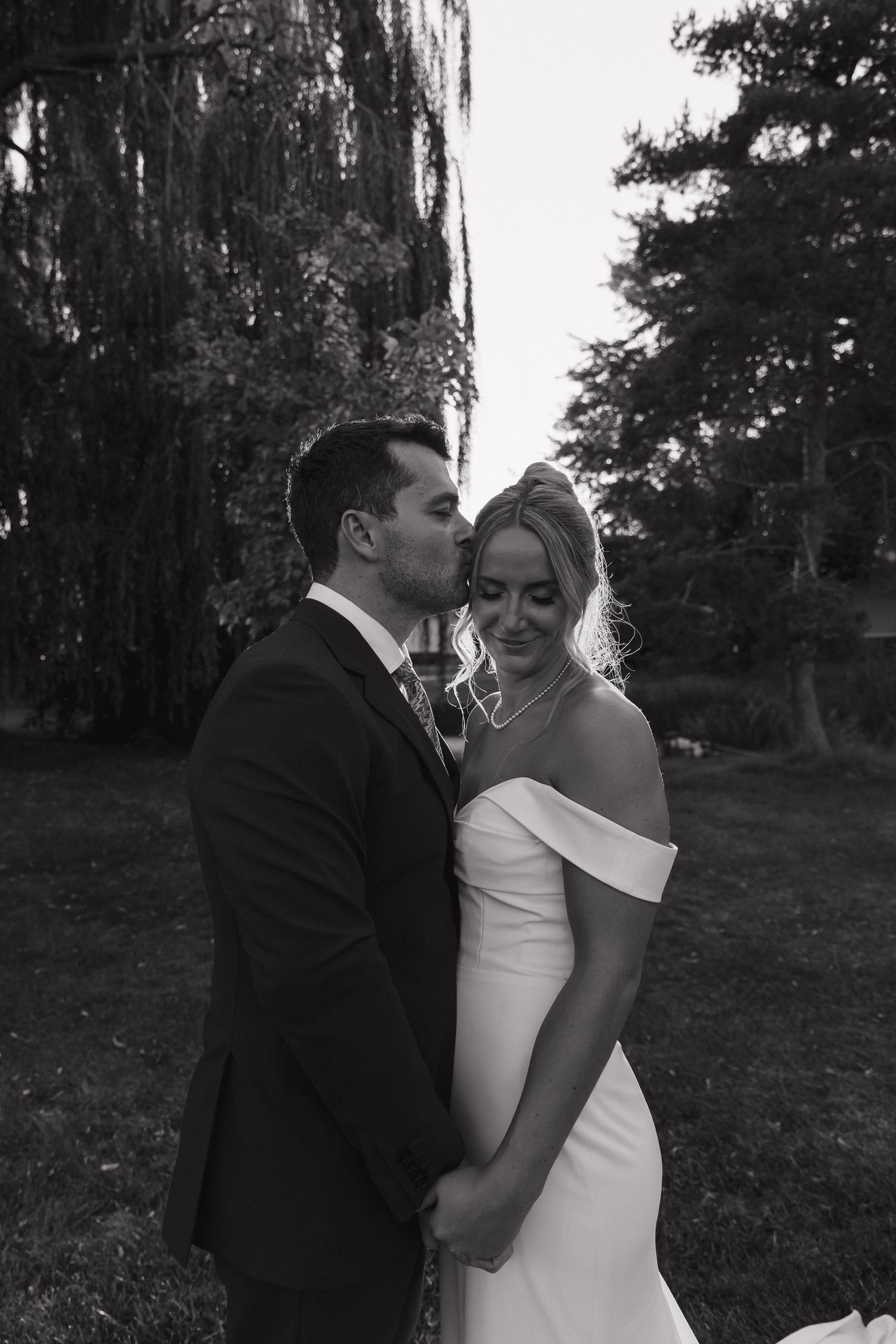 Black and white photo of groom kissing bride's cheek as she smiles in an outdoor garden setting.