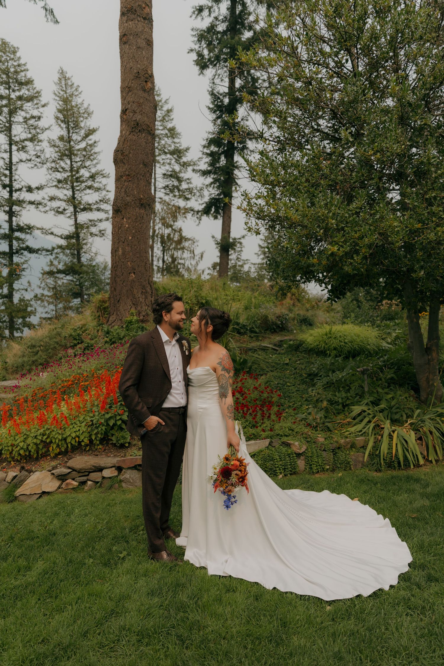 Bride and groom share romantic moment in lush garden with tall evergreen trees and red flowers.