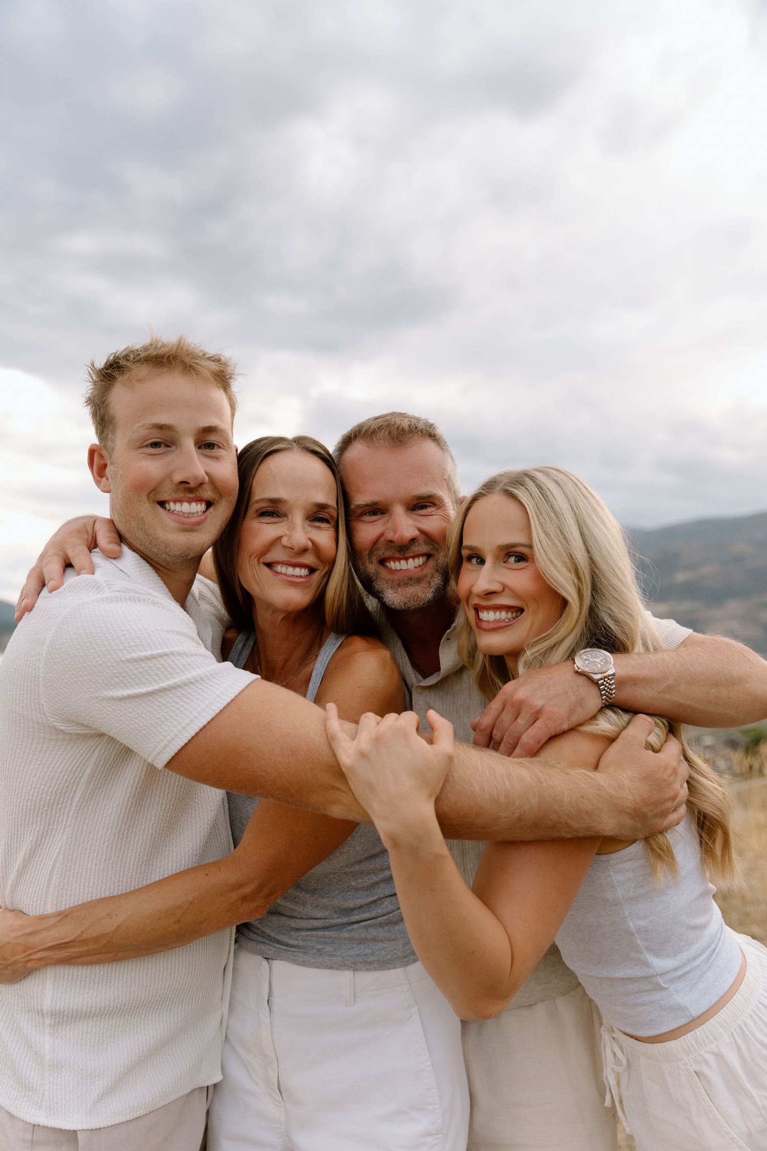 Happy family of four in white outfits hugging and smiling together outdoors under a cloudy sky.