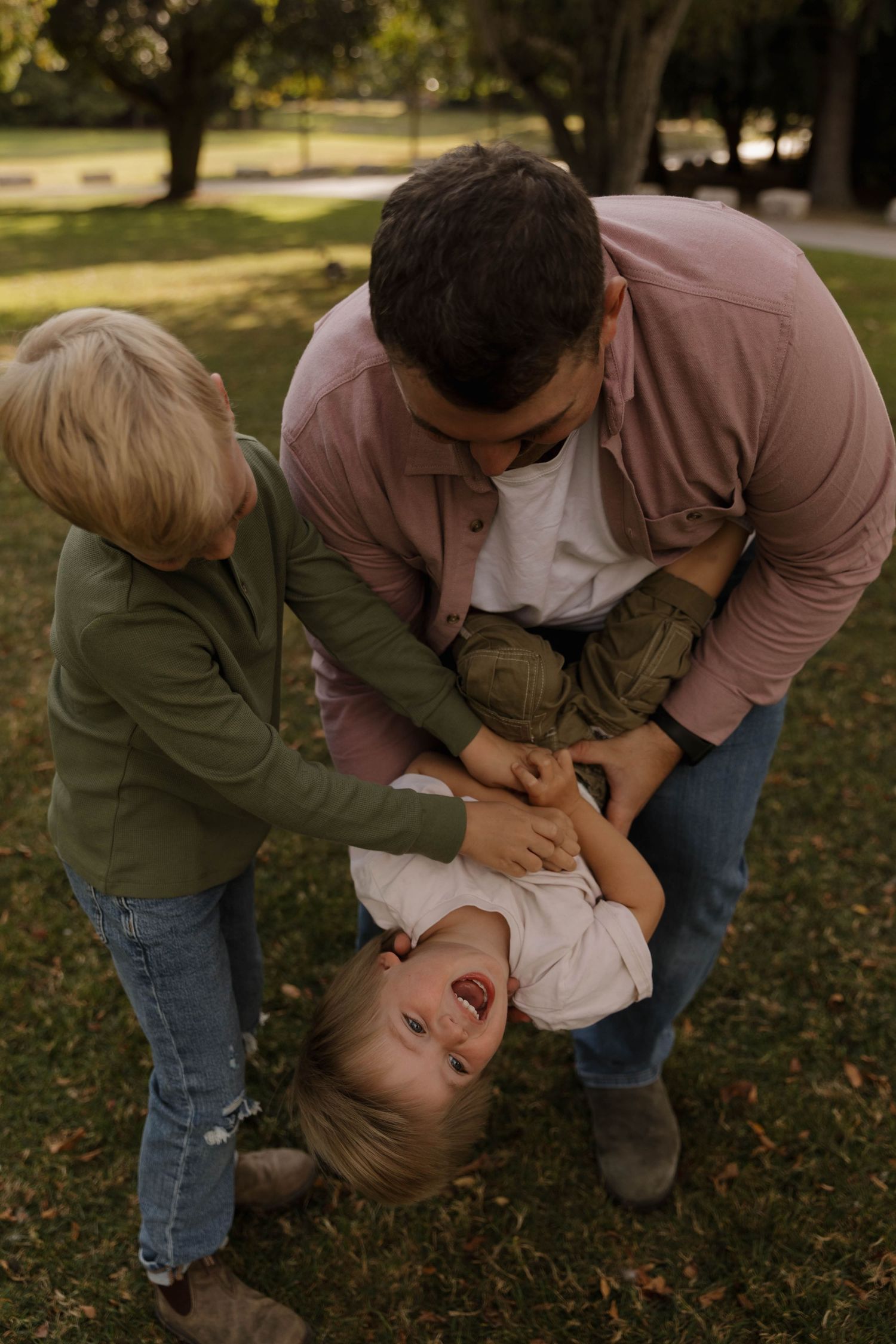 Father playfully tickling laughing child while sibling joins in at a park.
