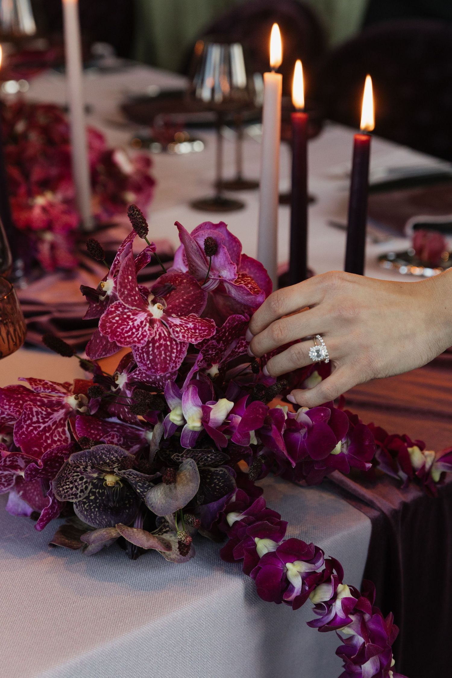 Brides hand with diamond ring rests on a lush burgundy and purple orchid table arrangement.