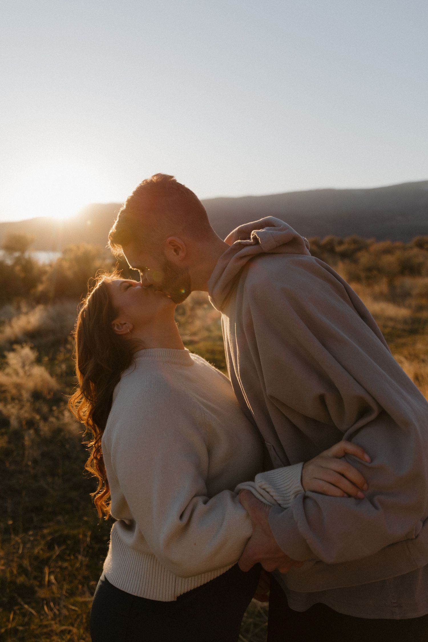 Couple sharing a romantic kiss at golden sunset in an open field with mountain backdrop.