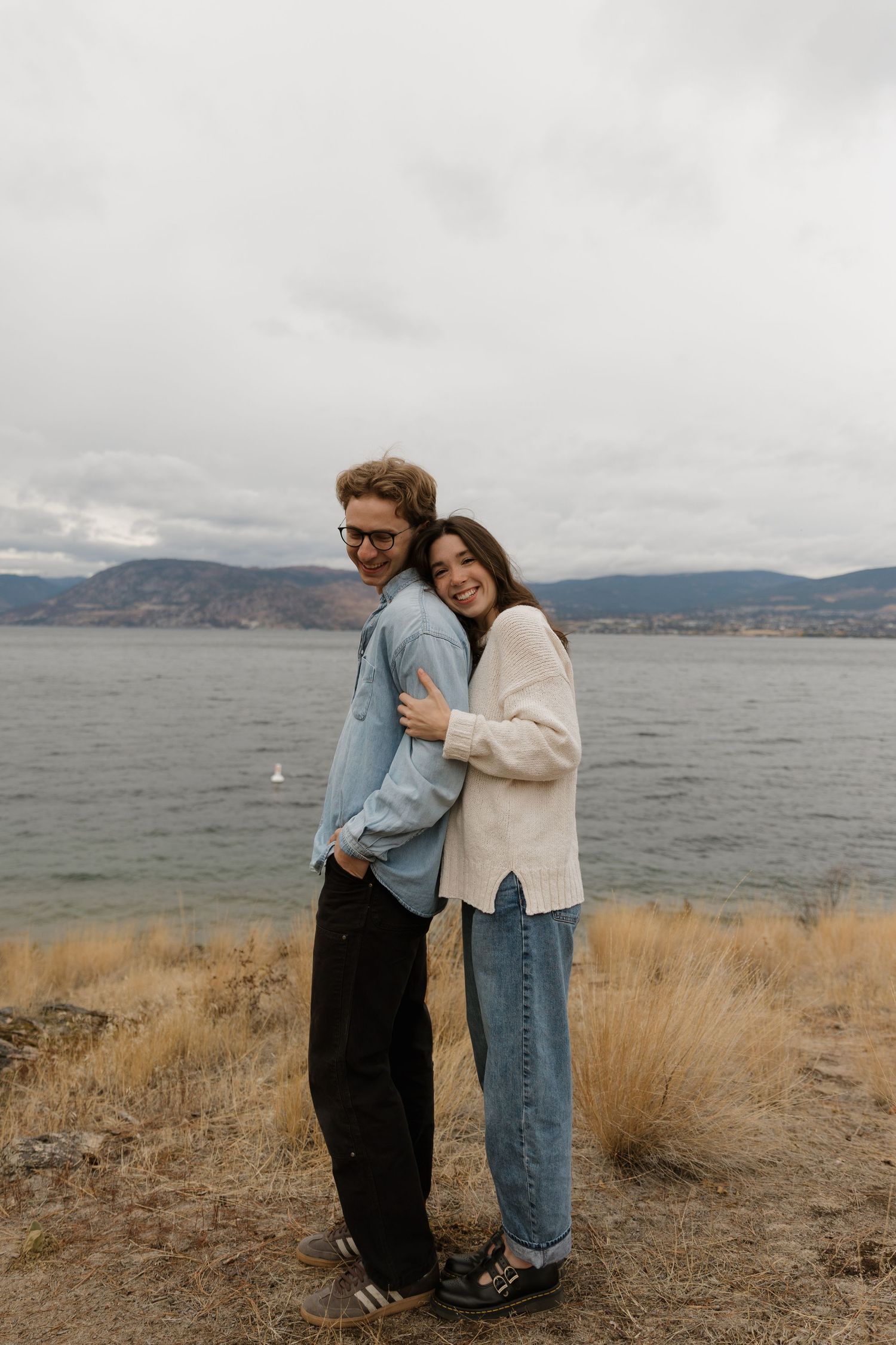 Smiling couple embracing back-to-back on a lakeside cliff with overcast mountain scenery.