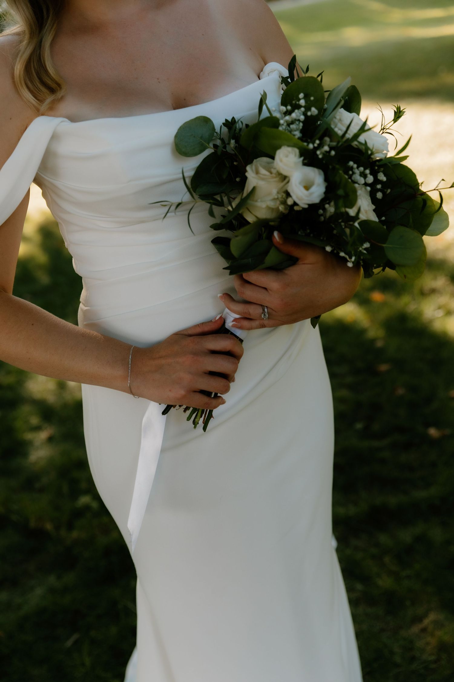 Bride in off-shoulder white gown holds elegant bouquet of white roses and greenery outdoors.