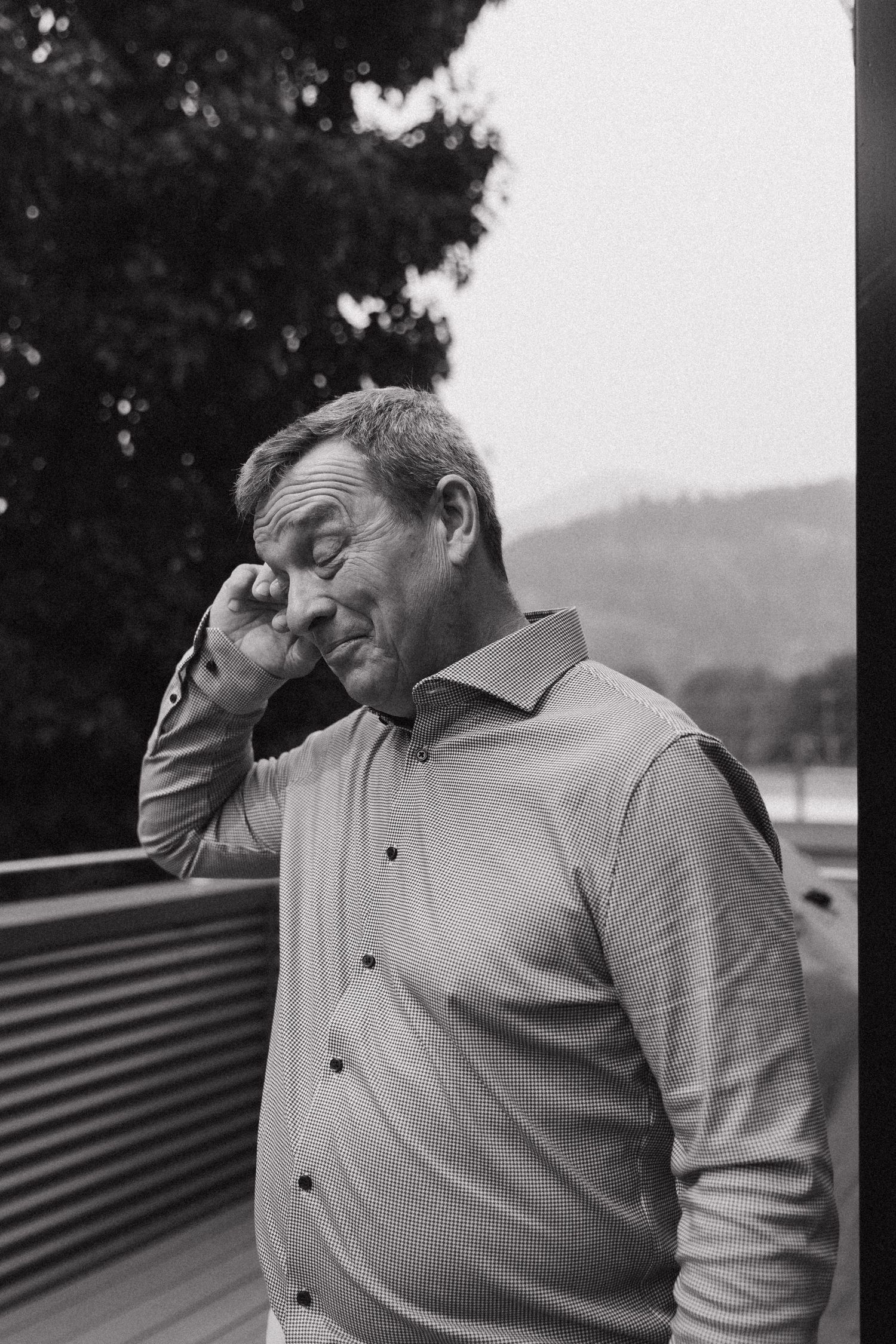 Black and white portrait of emotional older man wiping tear at outdoor wedding celebration.
