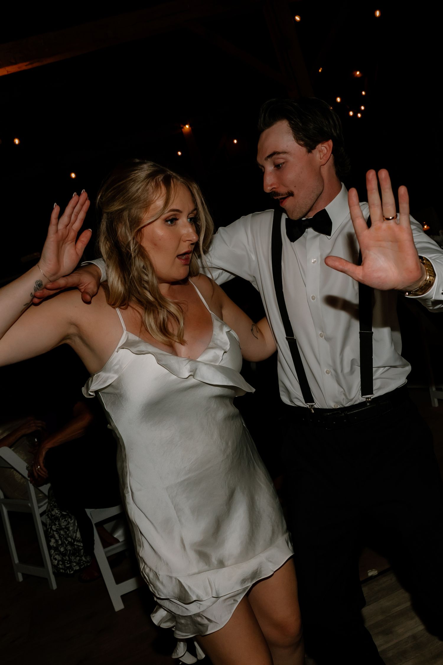 Bride and groom dancing energetically at wedding reception in rustic barn with warm ambient lighting.