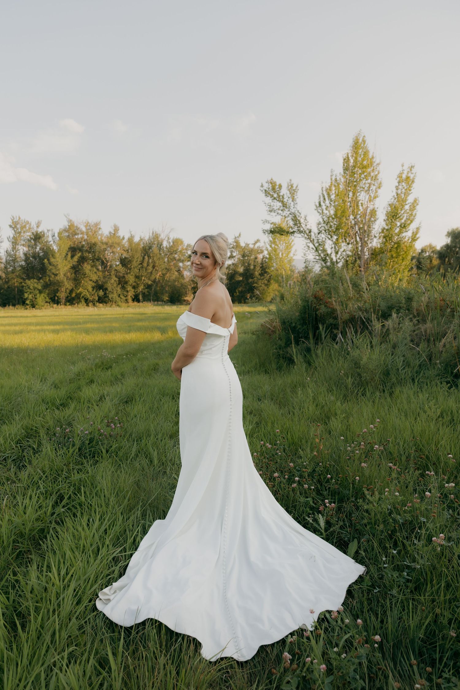 Bride in elegant off-shoulder white wedding gown with train poses in a sunlit green meadow.