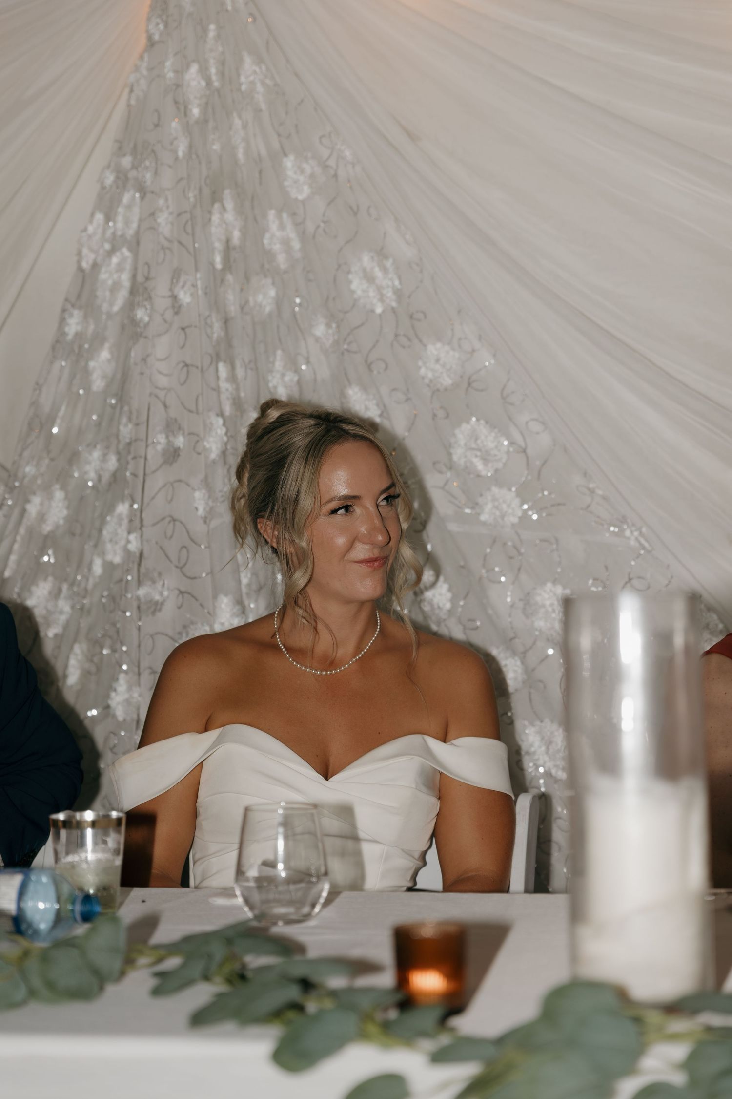 Bride in off-shoulder white gown smiling softly at wedding reception beneath floral draping.