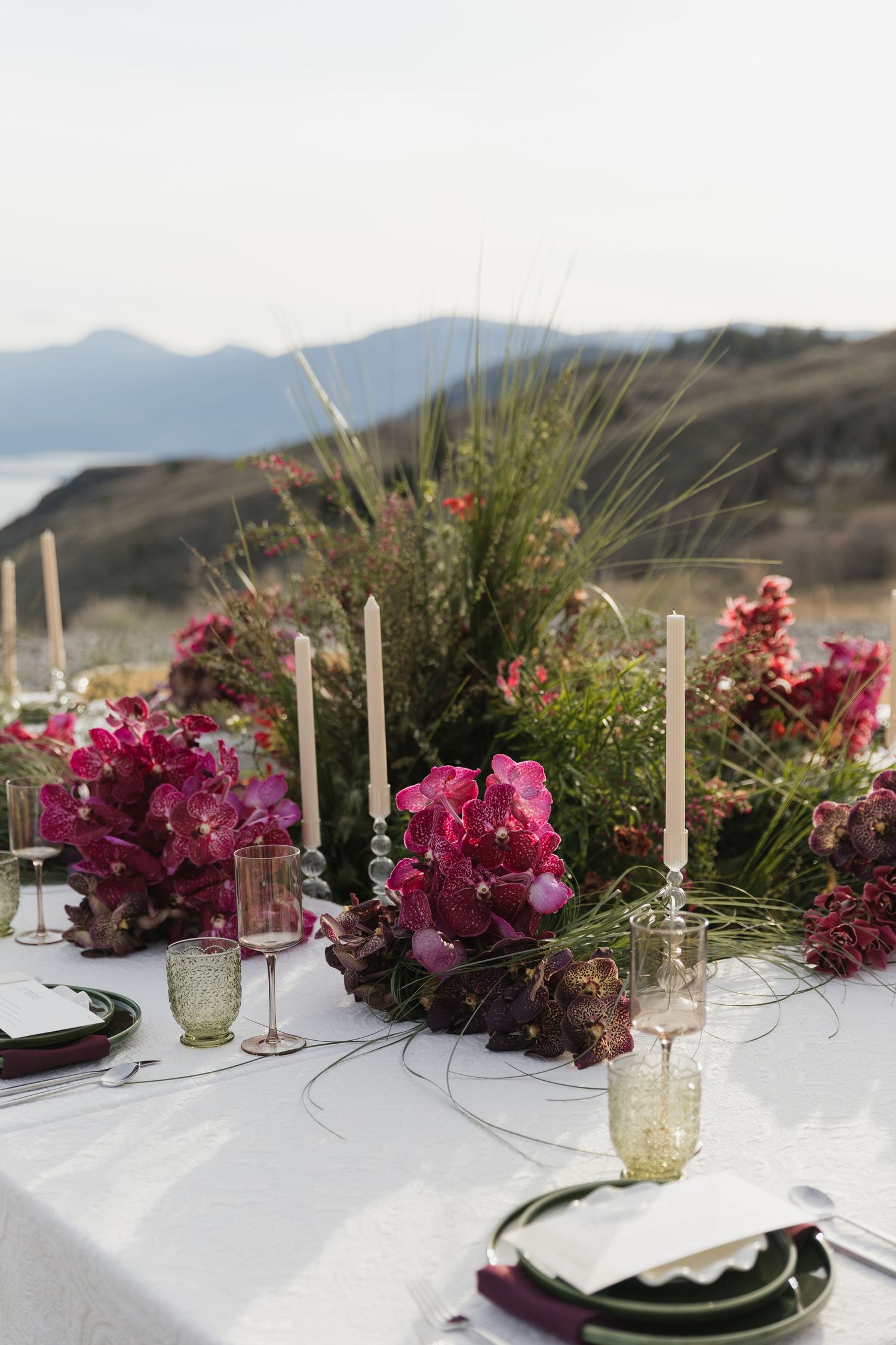 Elegant outdoor wedding table setting with pink orchids, tall ivory candles, and scenic coastal mountain backdrop.
