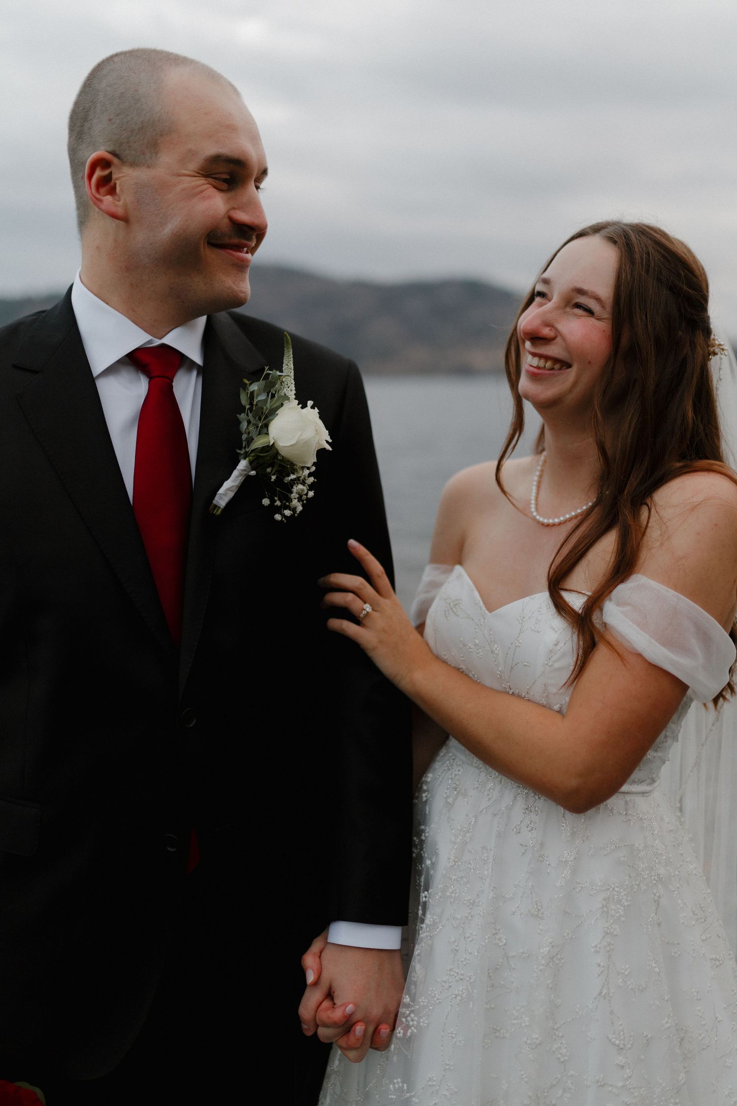 Smiling bride and groom holding hands outdoors with scenic lake and mountain backdrop on their wedding day.