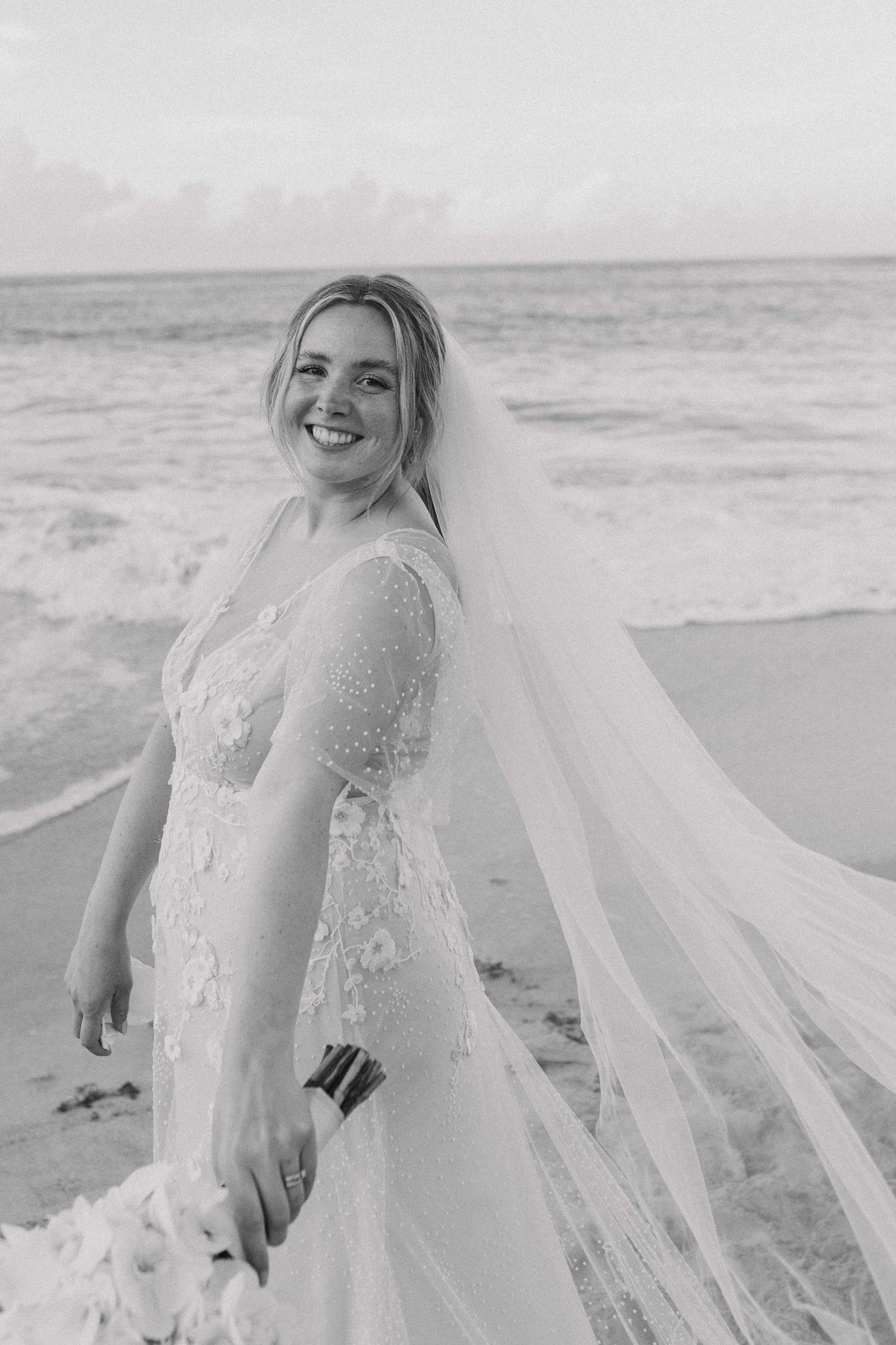 Black and white portrait of smiling bride in lace gown with flowing veil on beach.