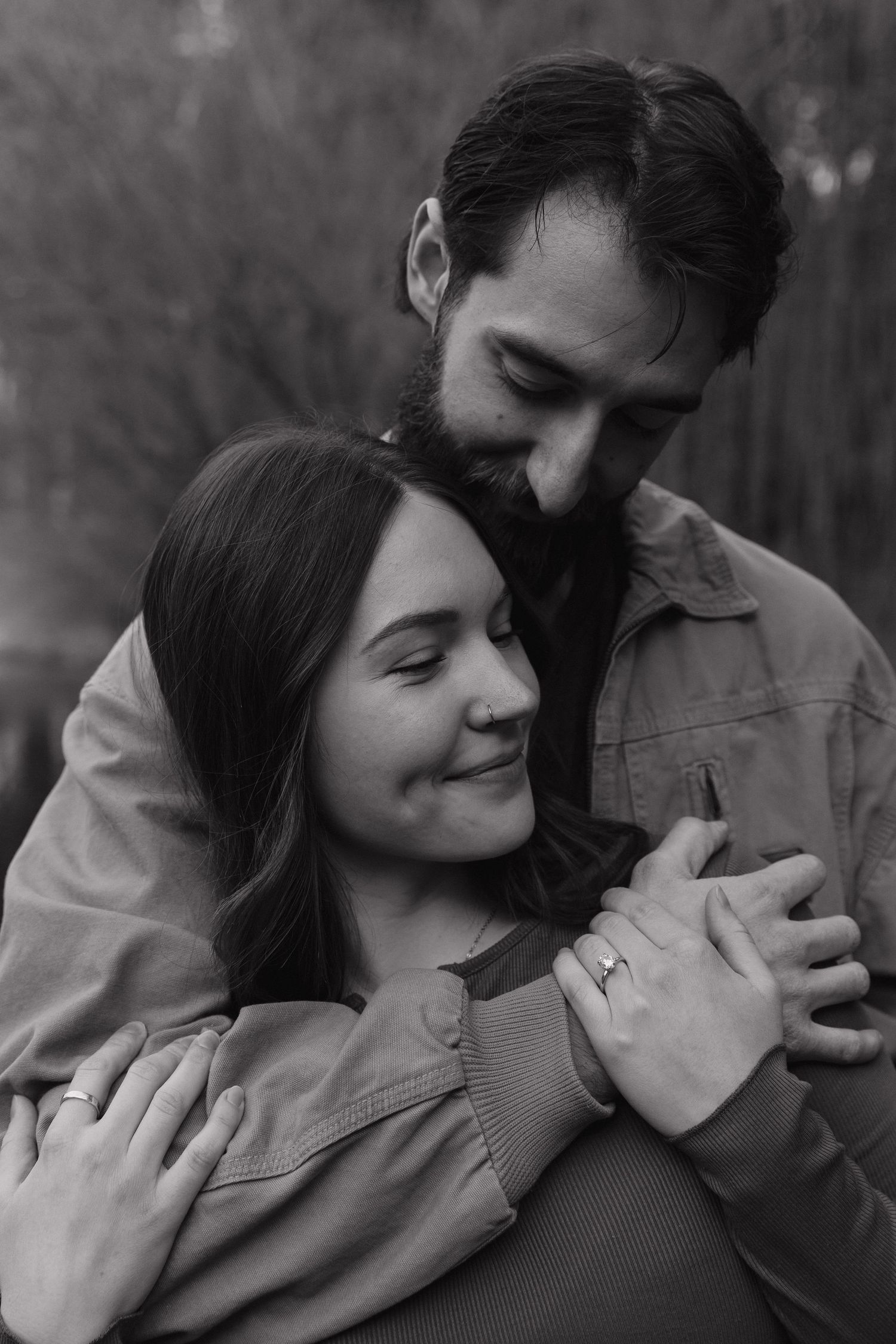 Black and white photo of a bearded man embracing a smiling woman from behind outdoors.