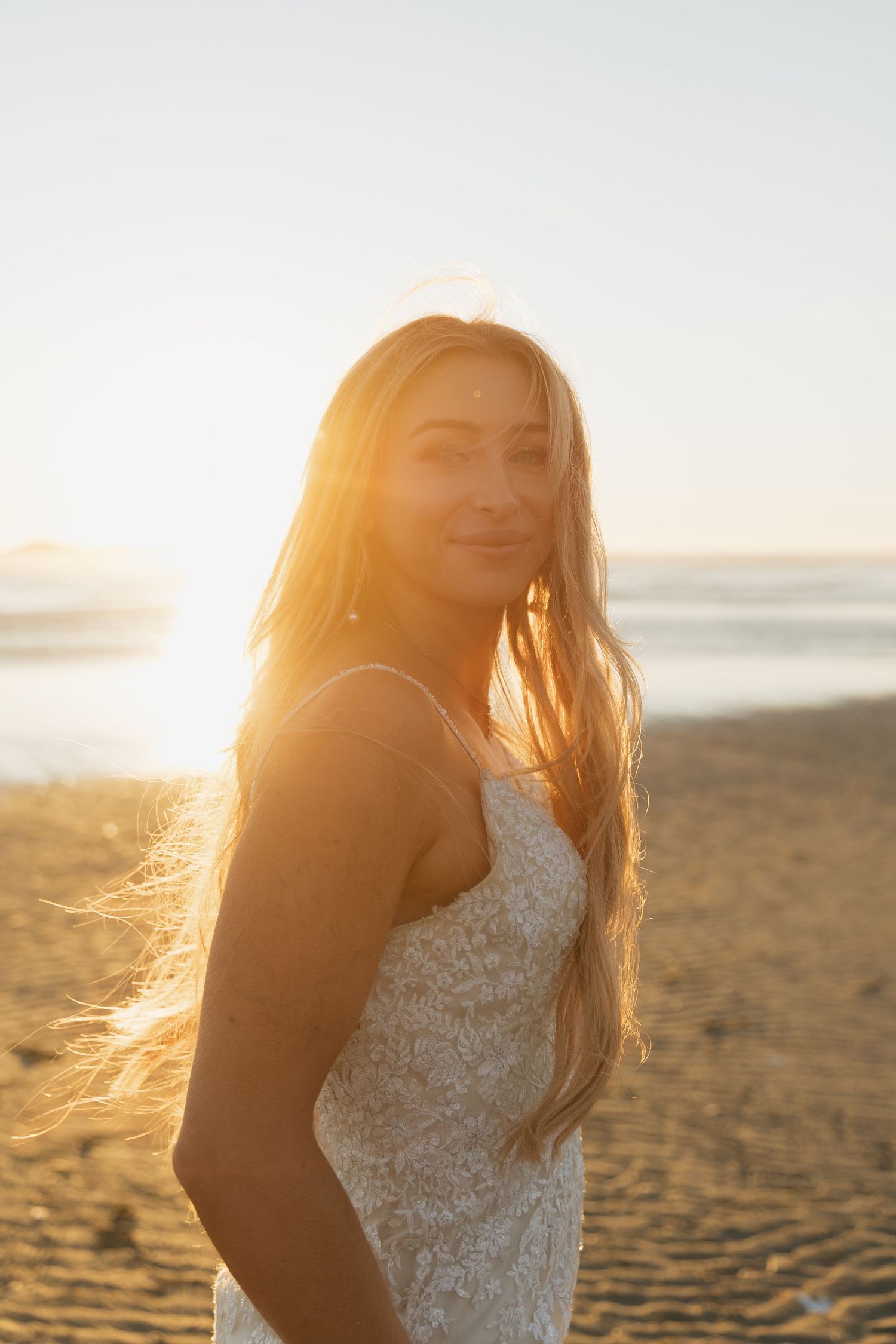 Radiant bride in lace gown stands on a sunlit beach, golden hour light illuminating her long hair.