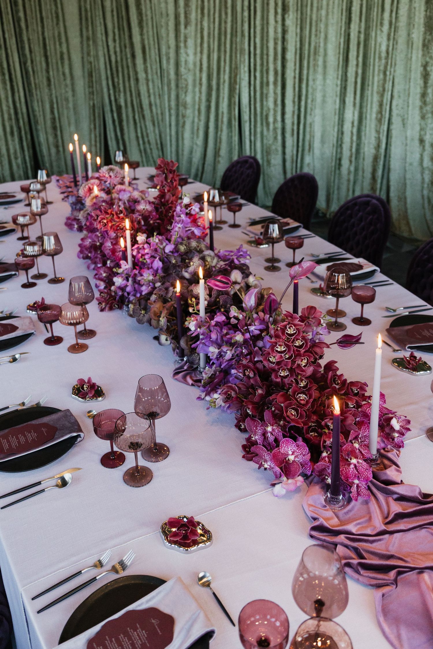 Elegant wedding reception table with purple and burgundy floral centerpieces, candles, and tinted glassware.