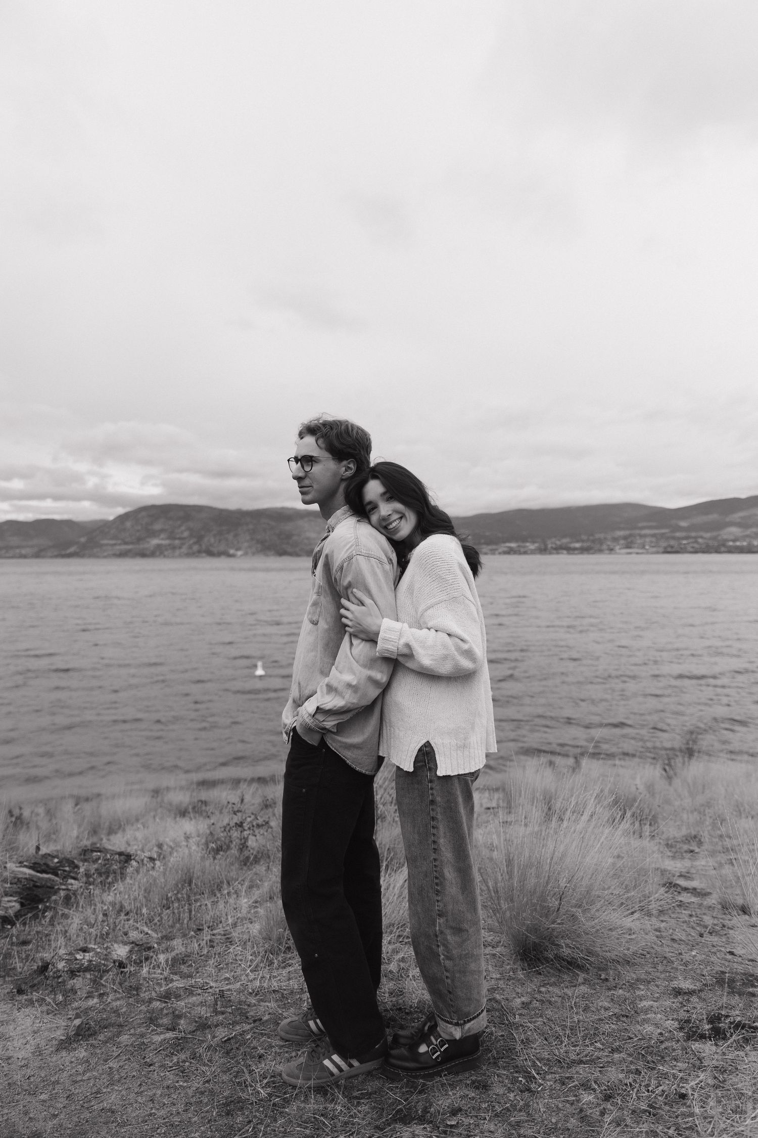 Black and white photo of couple embracing by a lake with hills in the background.