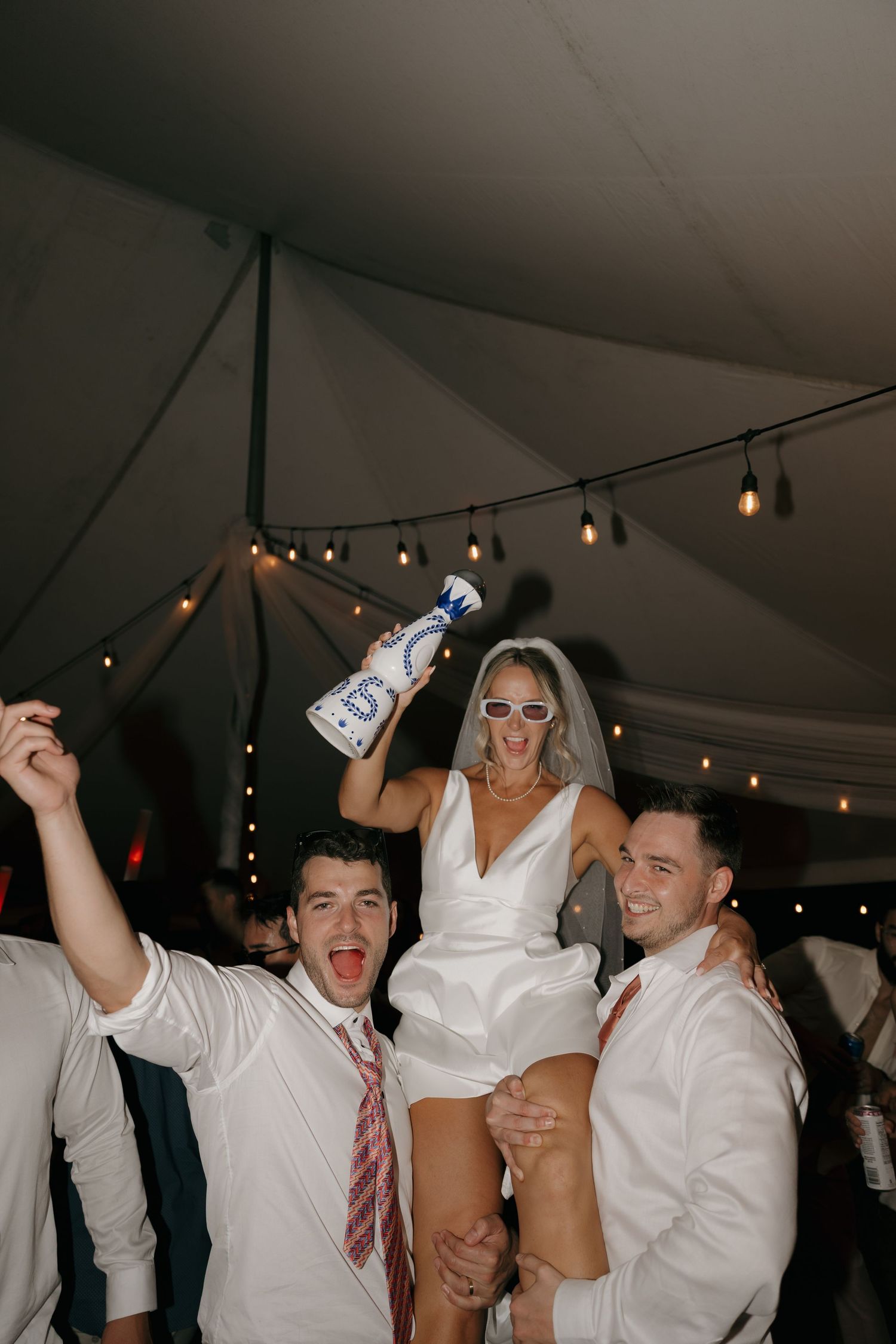 Bride in white mini dress holding tequila bottle on shoulders of groomsmen dancing at tented wedding reception.
