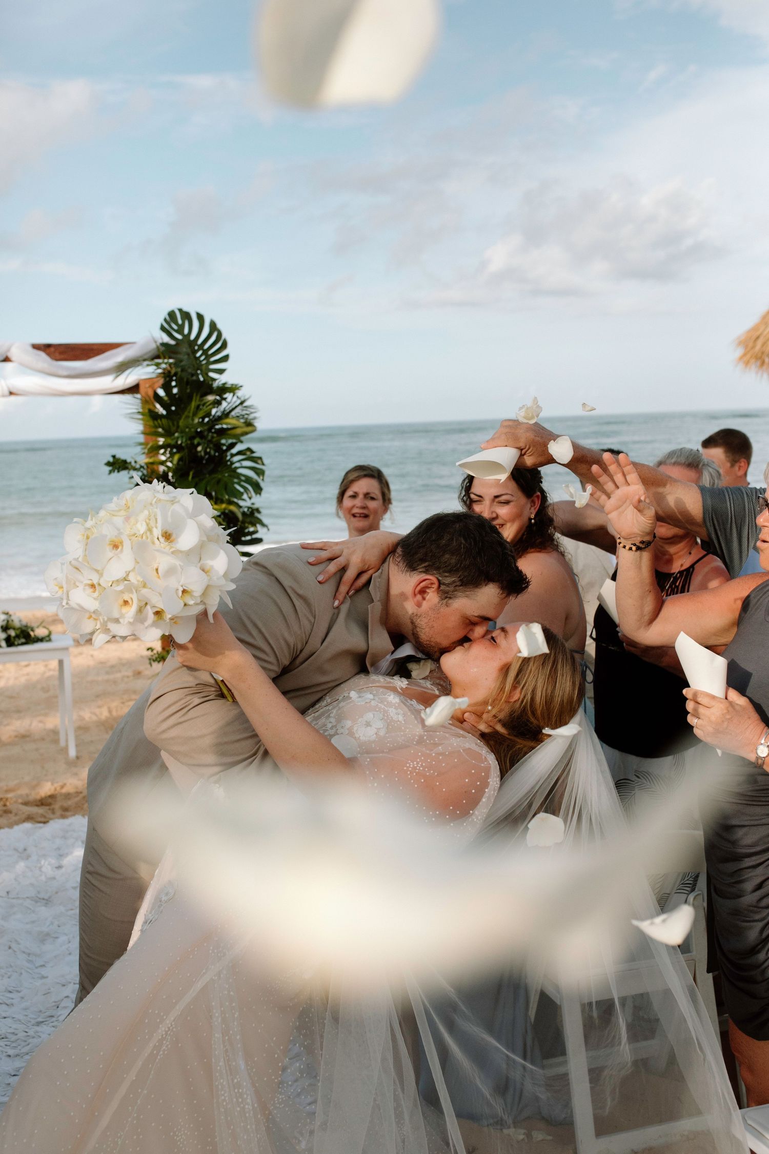 Bride and groom share first kiss surrounded by cheering guests at beach ceremony.