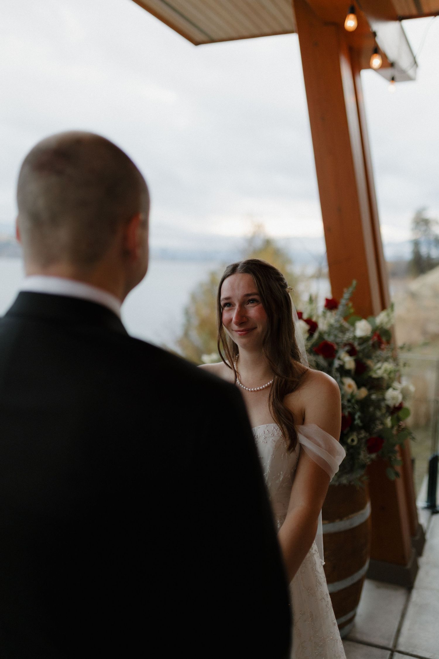 Radiant bride in off-shoulder lace gown smiling at groom during intimate lakeside wedding ceremony.