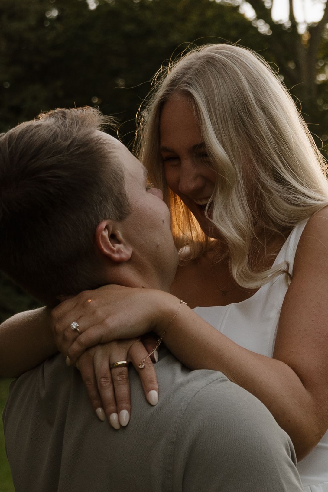 A smiling blonde woman in a white dress being lifted by her partner in a lush green park.