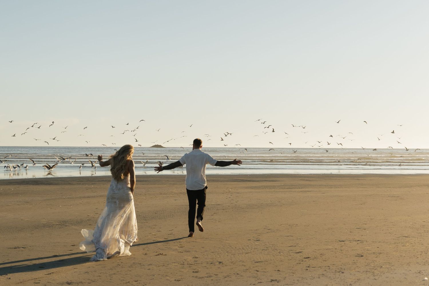 Bride and groom run joyfully along a sandy beach at sunset with seagulls soaring overhead.