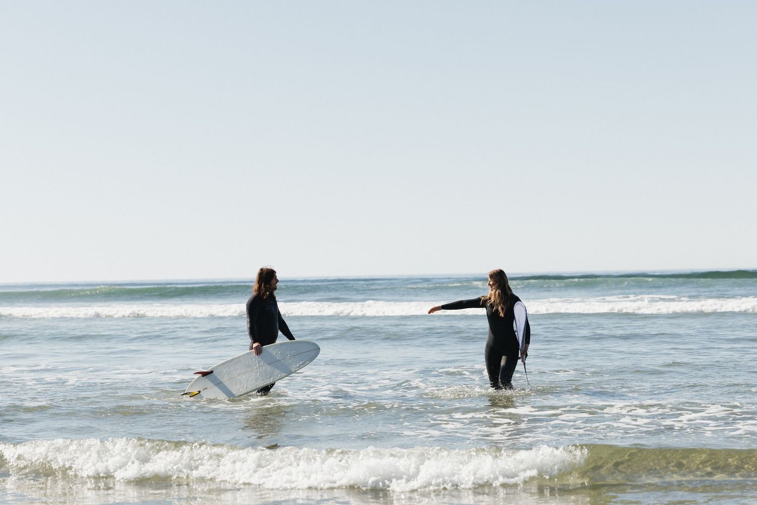 Two surfers in wetsuits walking into ocean waves on a bright sunny day at the beach.