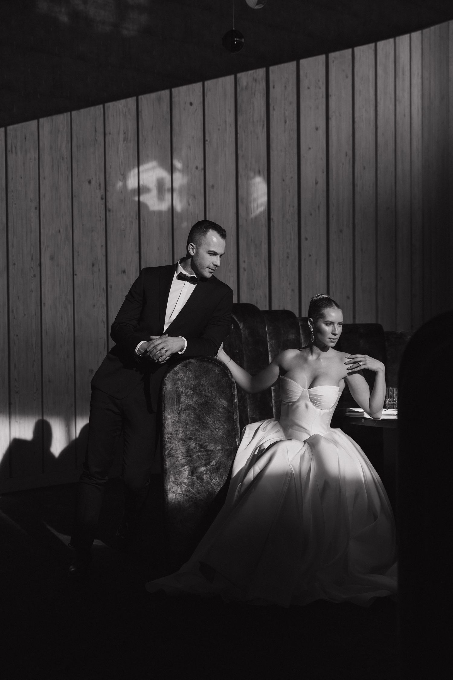 Black and white photo of groom in tuxedo standing behind bride in strapless ball gown at wedding reception.