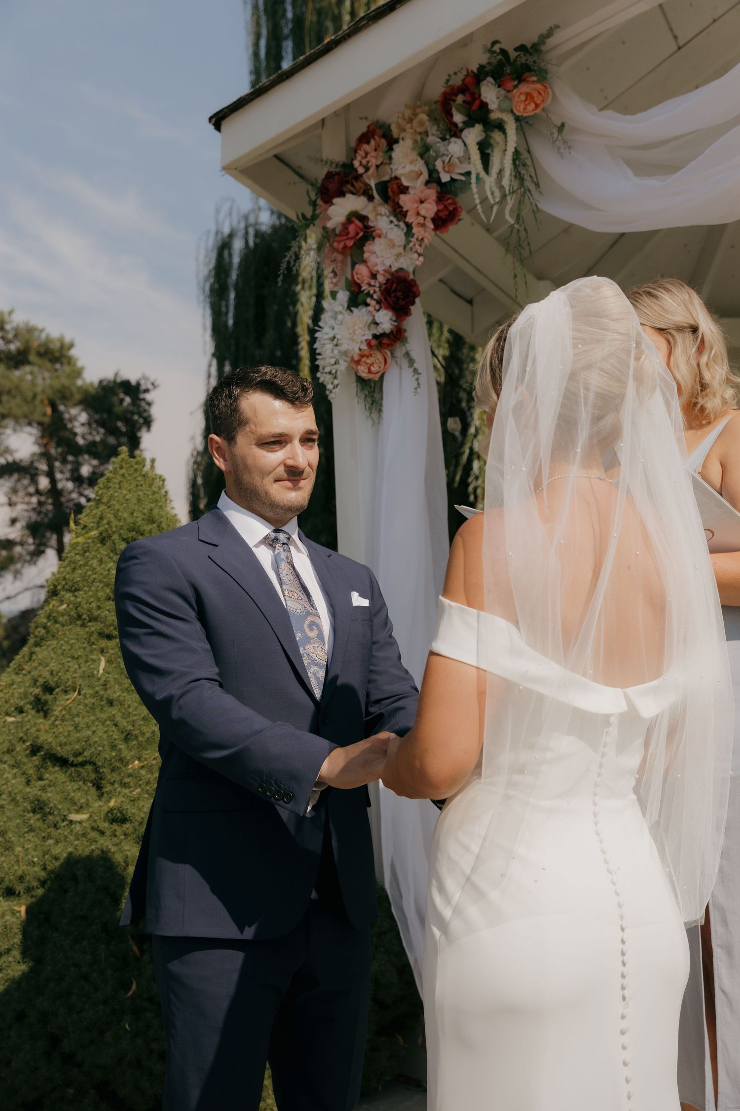Groom in navy suit smiling at bride during outdoor wedding ceremony at floral altar.