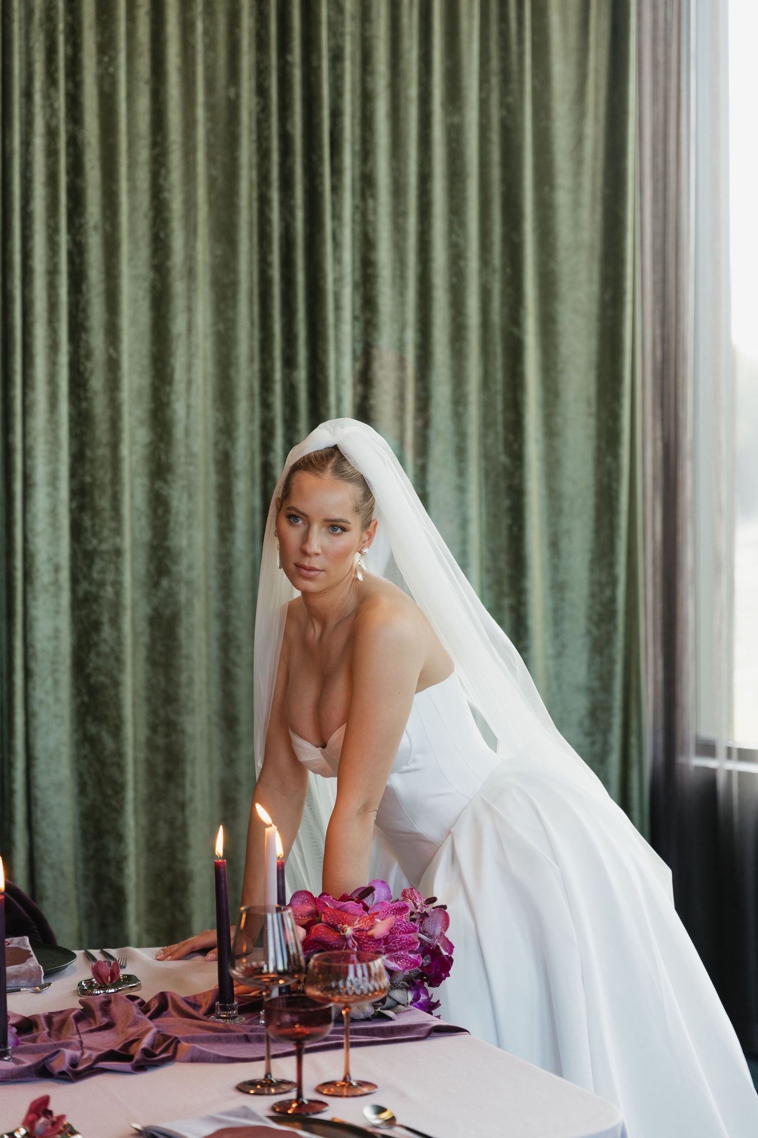 Elegant bride in white strapless gown and veil leans over candlelit wedding table adorned with purple orchids.