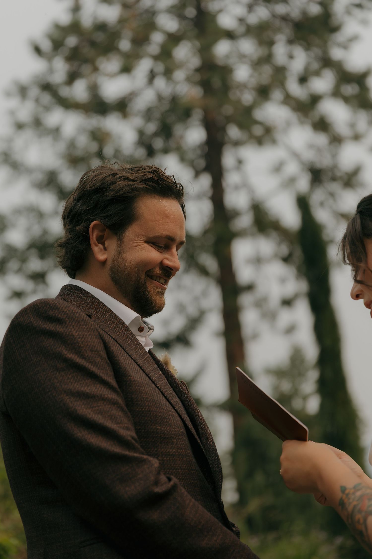 Smiling groom in brown suit reading wedding vows during an outdoor elopement ceremony.