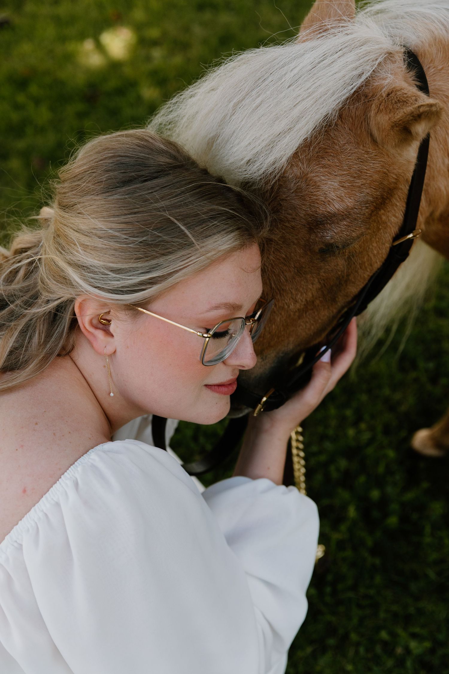 Blonde bride in glasses gently rests her head against a brown horse with a white mane.