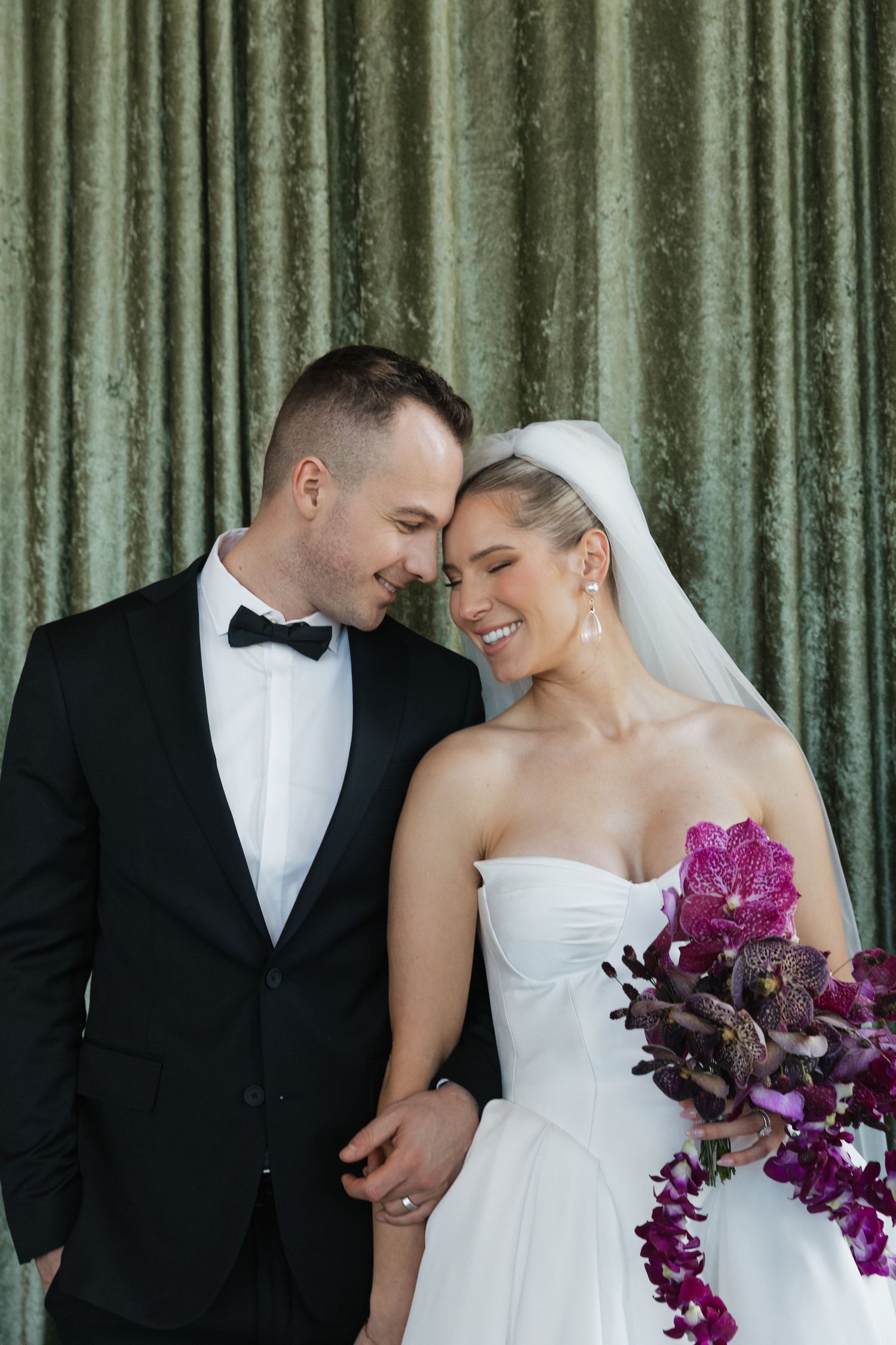Smiling bride and groom touch foreheads tenderly, bride holding cascading purple orchid bouquet before green velvet backdrop.