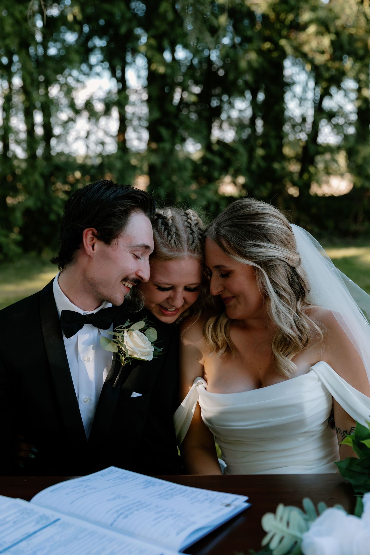 Bride, groom in black tuxedo, and officiant sharing joyful moment during outdoor wedding signing ceremony.