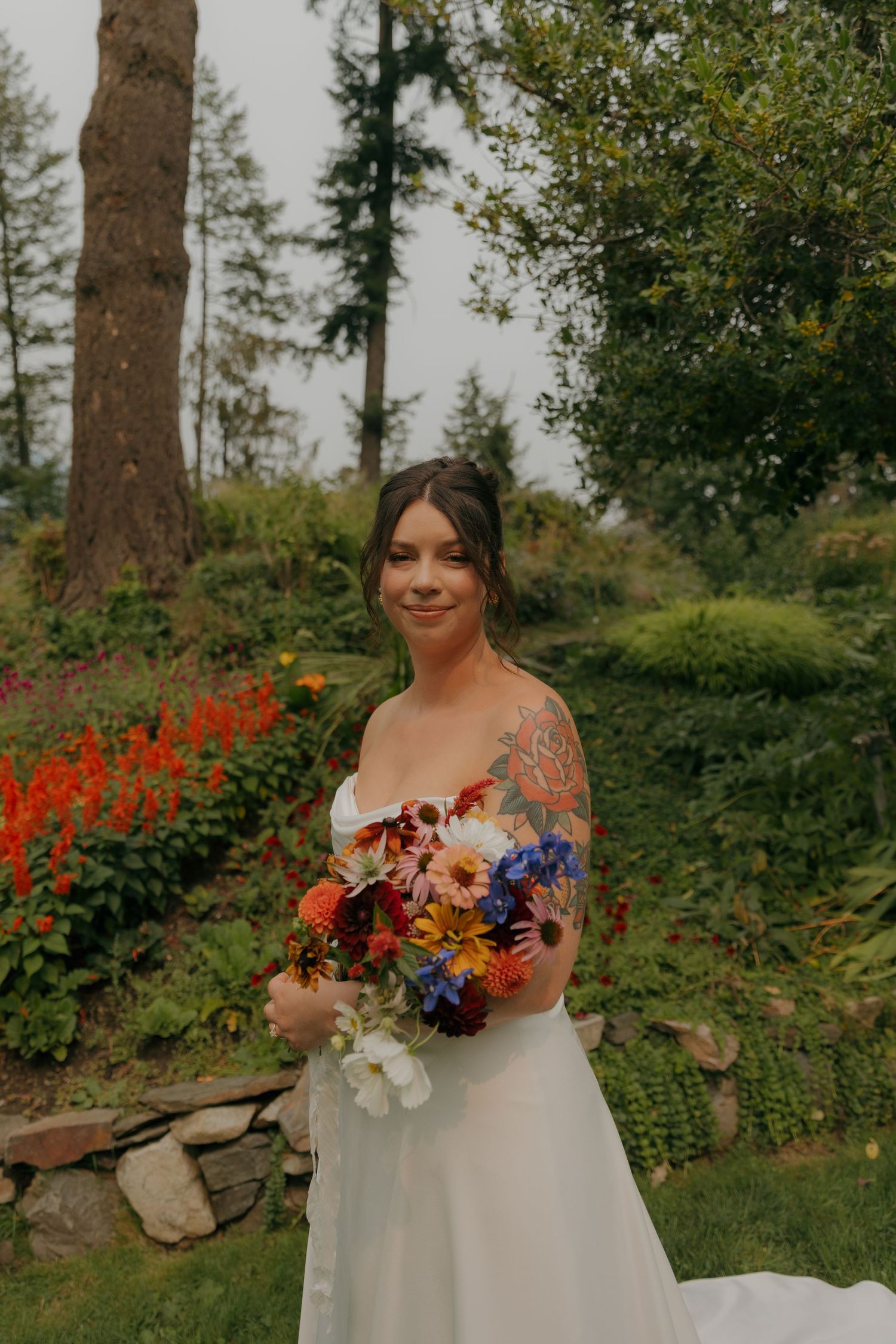 Tattooed bride in strapless white gown smiles holding vibrant wildflower bouquet in garden.