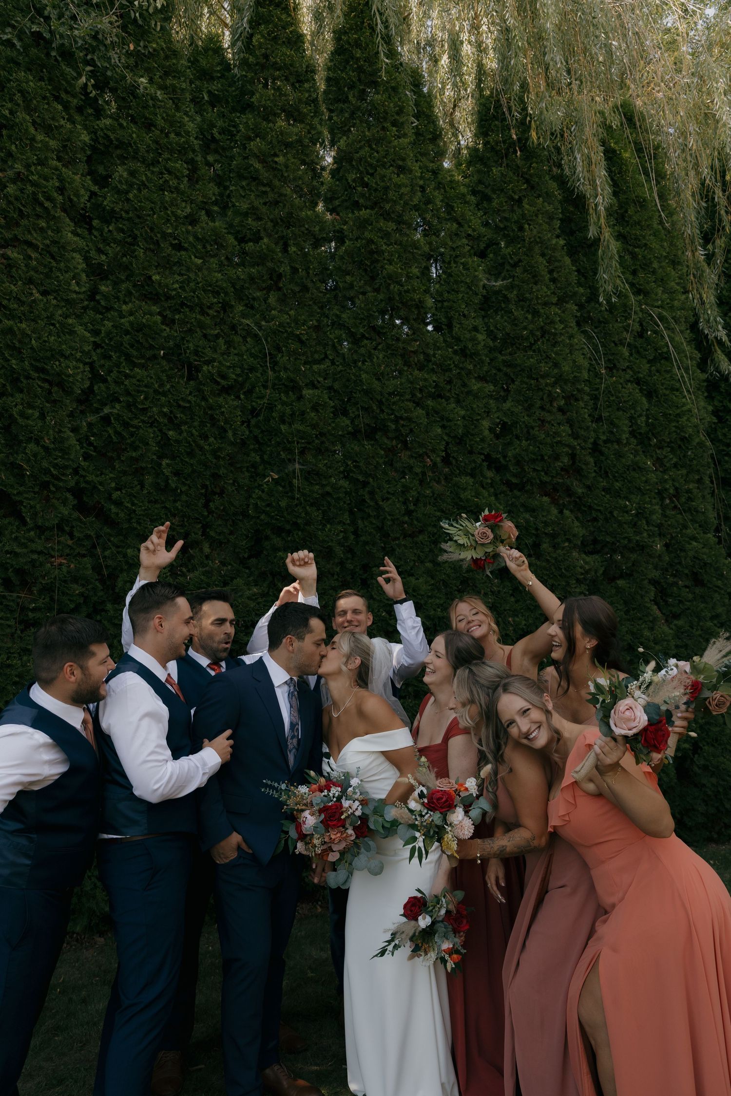 Wedding party celebrating outdoors with bridesmaids in coral dresses and groomsmen in navy suits holding bouquets.
