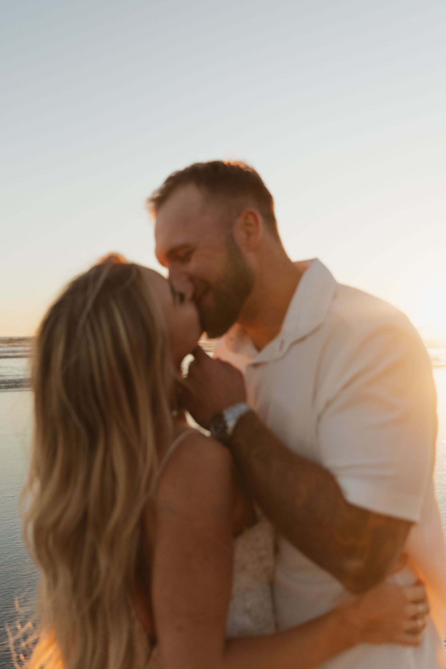 Couple embracing and smiling at sunset on a beach, man with tattoos in white shirt holding blonde woman.