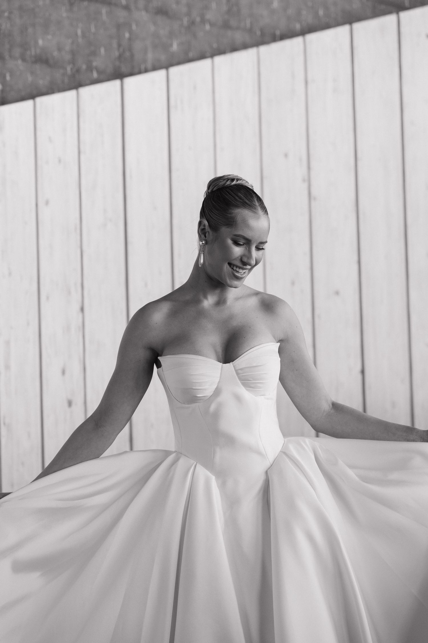 Black and white photo of smiling bride in strapless ballgown twirling near wooden wall backdrop.