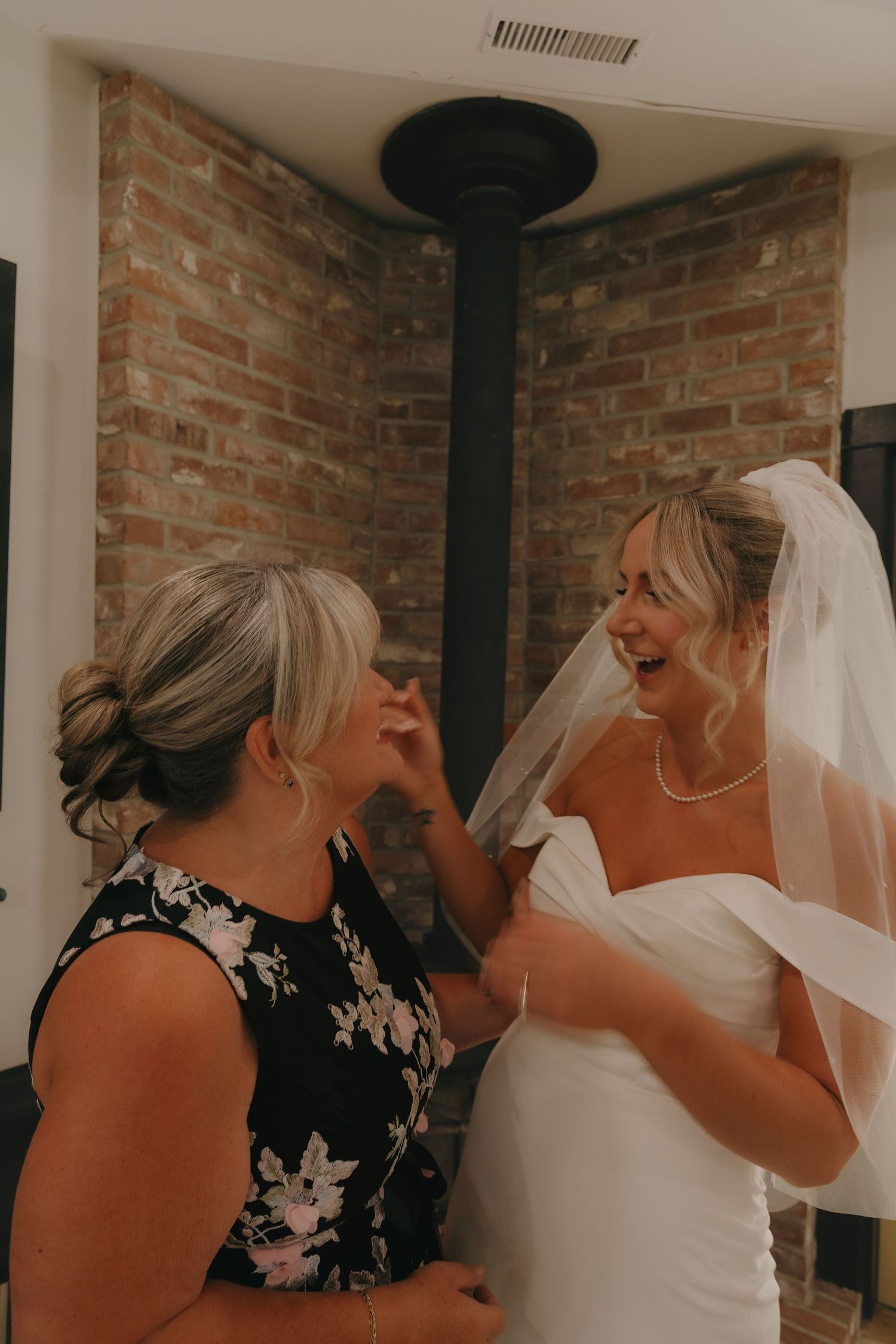 Laughing bride in white gown and veil shares a joyful moment with a woman indoors near brick wall.