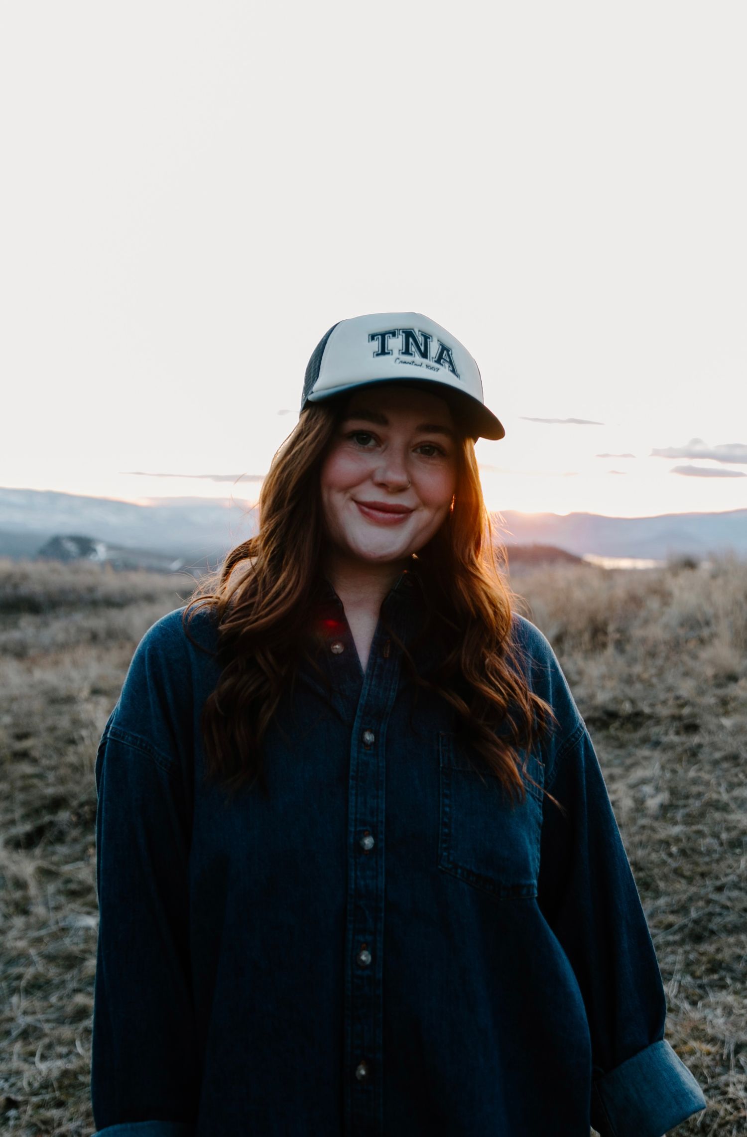 Smiling woman wearing a TNA trucker hat at golden hour in a field.